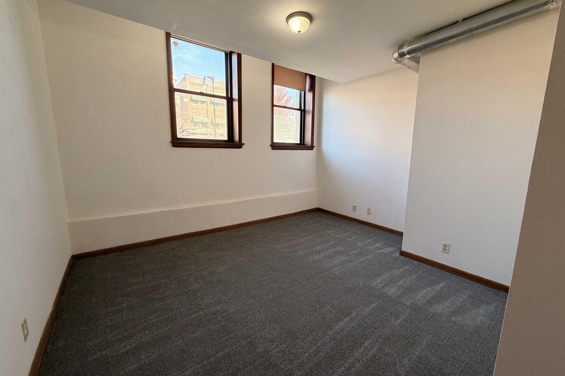 A vacant room with light gray carpeted flooring, two windows allowing natural light, and white walls. One wall features a metal duct. The room appears clean and spacious, suitable for various uses.