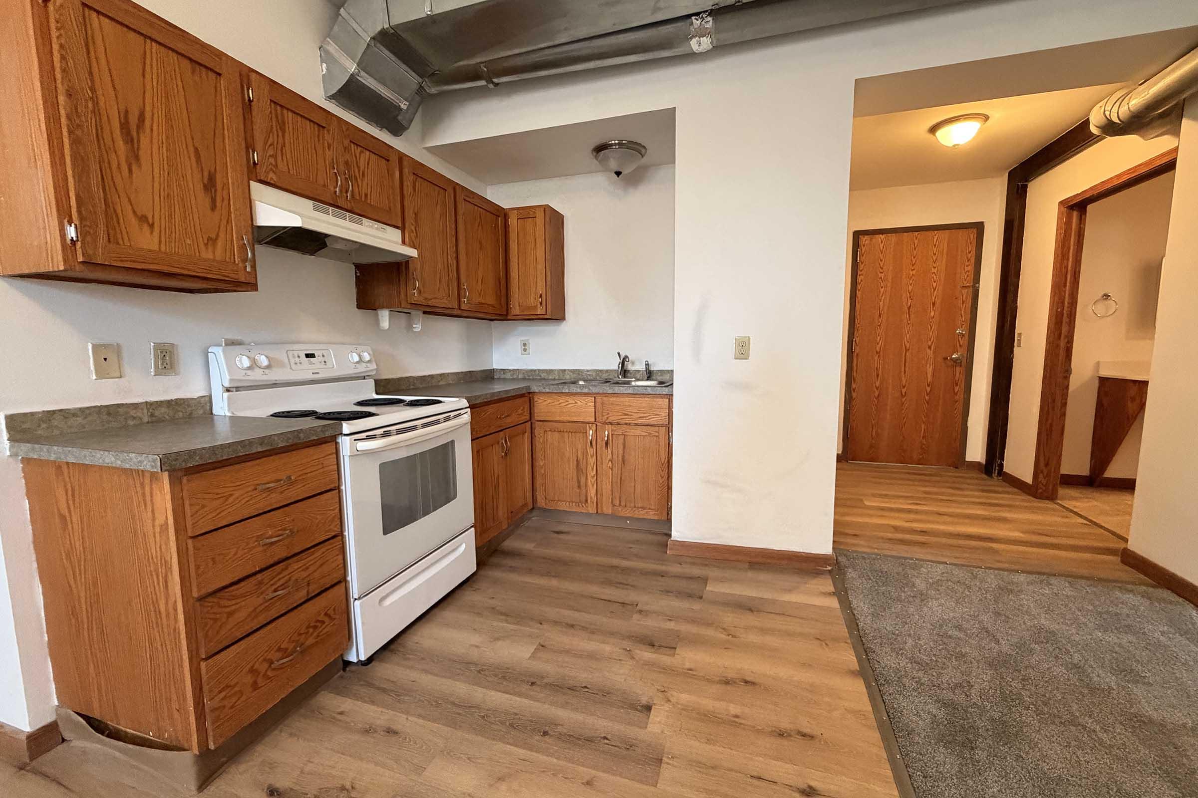 A view of a kitchen featuring wooden cabinets, a white stove, and a countertop. The room has a door leading to another space and visible flooring that transitions from hardwood to carpet. Natural light is present, highlighting the layout and appliances.