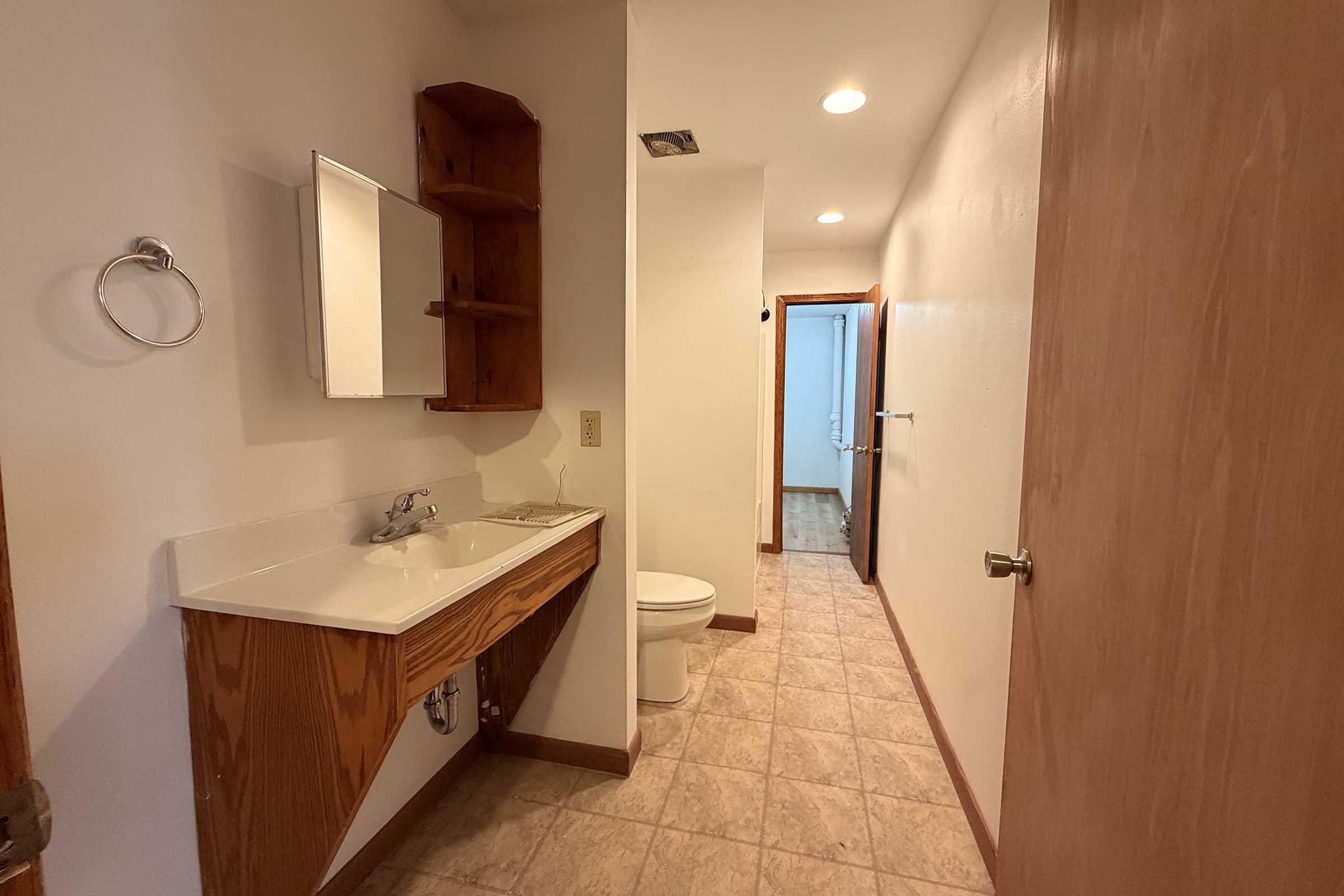 Interior view of a bathroom featuring a white sink with wooden storage below, a wall-mounted mirror, a toilet, and tiled flooring. The door is open, revealing a glimpse of a second room beyond. The walls are painted white, and there is a towel ring next to the sink.