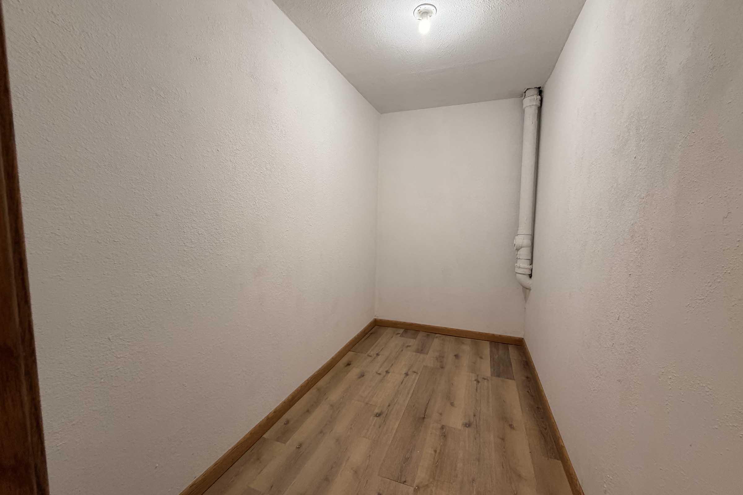 A small, empty room with plain white walls and a wooden floor. A light bulb hangs from the ceiling, and a pipe runs along one wall. The space appears unused and minimalistic, with no furniture or decorations.