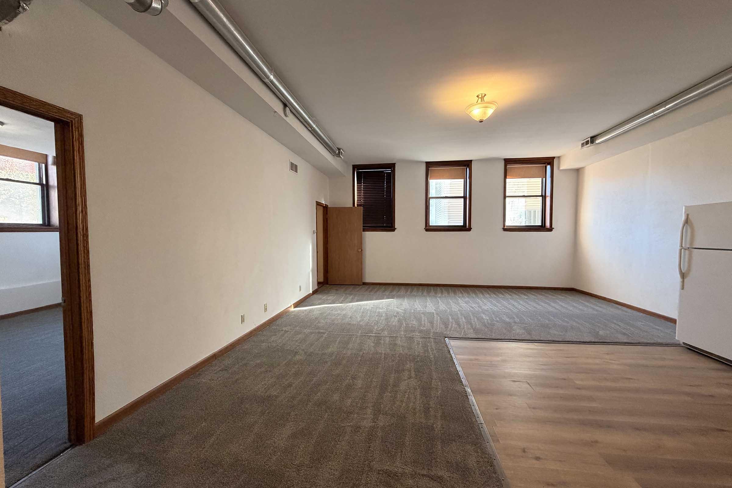 A spacious, empty room with light-colored walls, large windows with blinds, and a combination of carpet and hardwood flooring. A refrigerator is visible in the corner, and there are light fixtures on the ceiling. The room appears to have been recently cleaned and is ready for occupancy.
