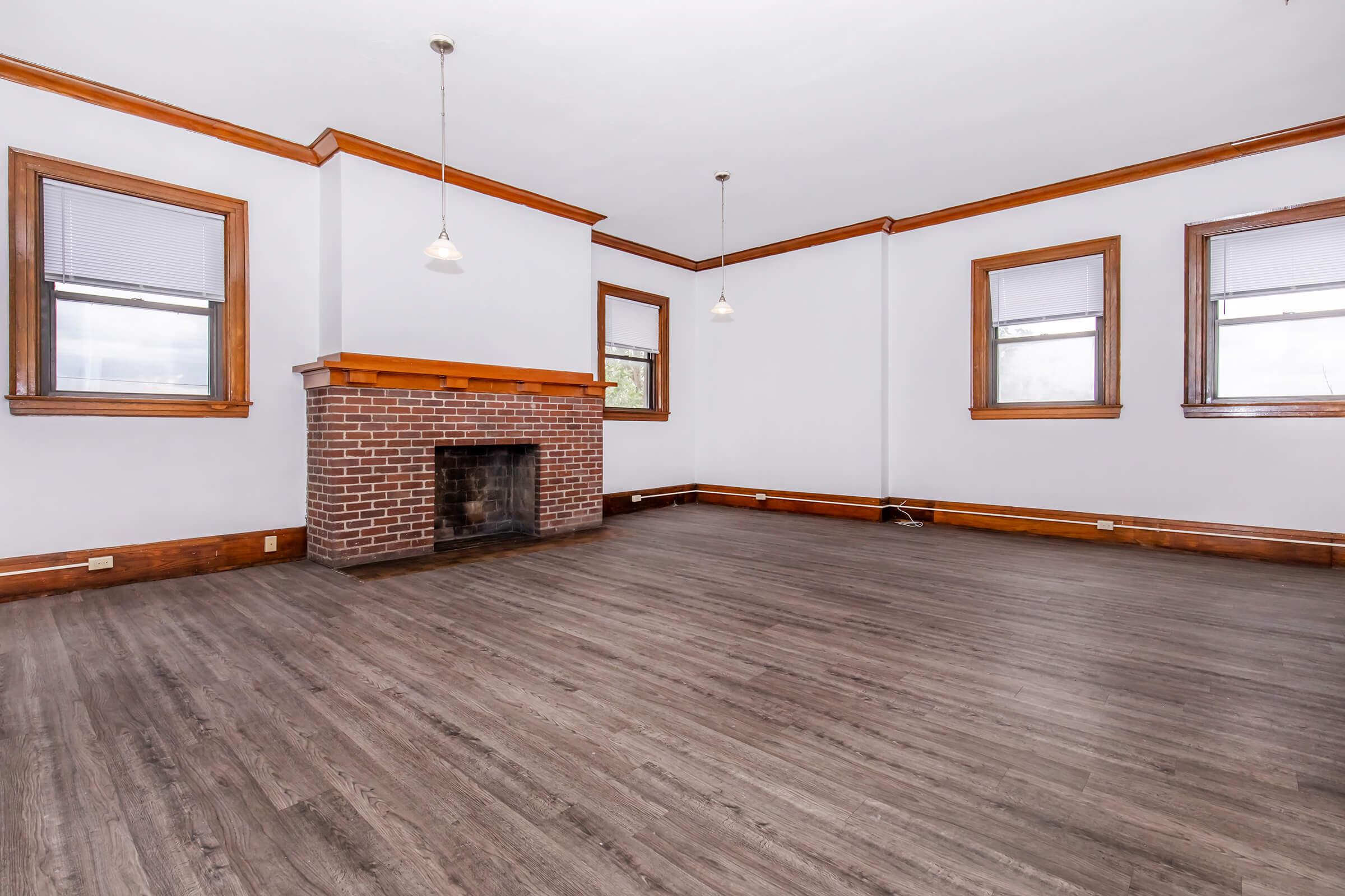 A spacious, empty living room featuring hardwood flooring, a brick fireplace, and large windows with wooden trim. The walls are painted light gray, and there are two pendant lights hanging from the ceiling, creating a bright and airy atmosphere.