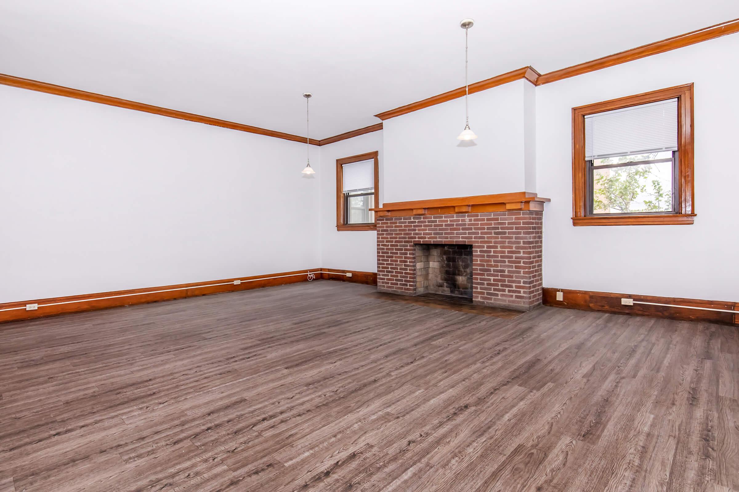 Spacious empty room with hardwood floors and exposed brick fireplace. The walls are painted light gray, and there are two windows with simple blinds. Decorative wood trim lines the room, and pendant lights hang from the ceiling, creating a bright and open atmosphere.