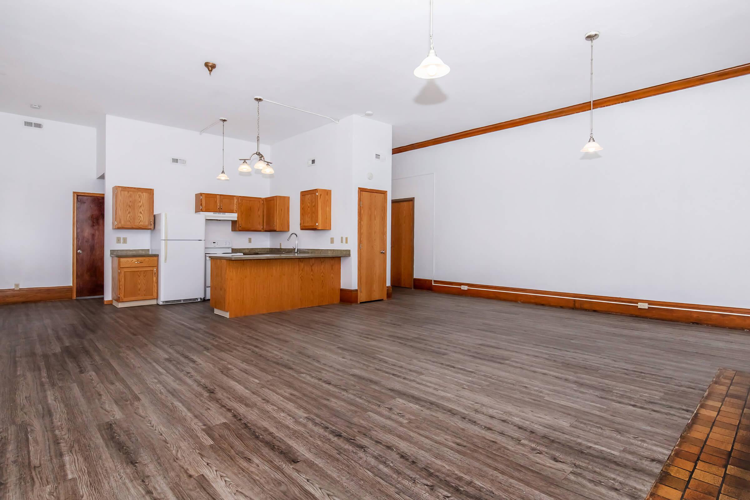 A spacious, empty interior of a residential apartment featuring wooden flooring, white walls, and a kitchen area with wooden cabinets, a refrigerator, and a stove. There are pendant lights hanging from the ceiling and no furniture is present, giving a clean and open feel to the space.