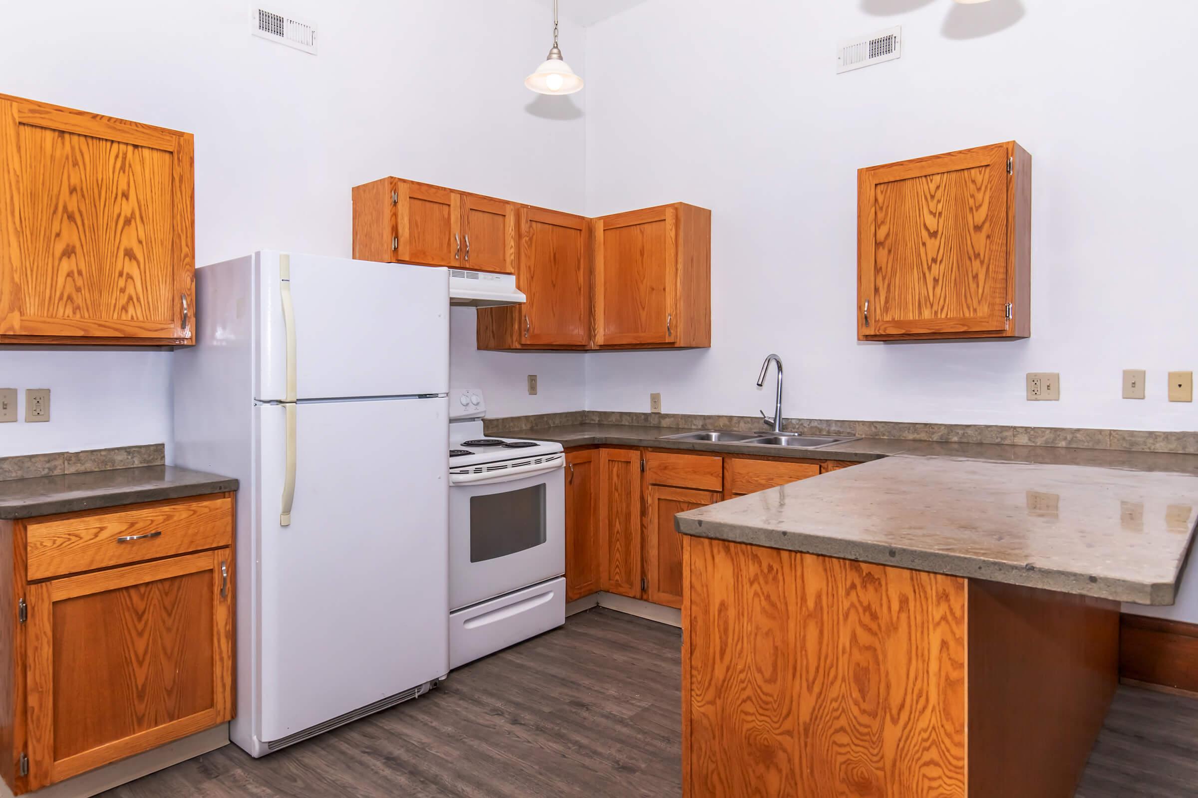 A kitchen with wooden cabinets and a countertop. It features a white refrigerator, a white stove with an oven, and a stainless steel sink. The floor has a dark, wood-like finish, and there is a light fixture hanging from the ceiling. The space is bright and well-organized.