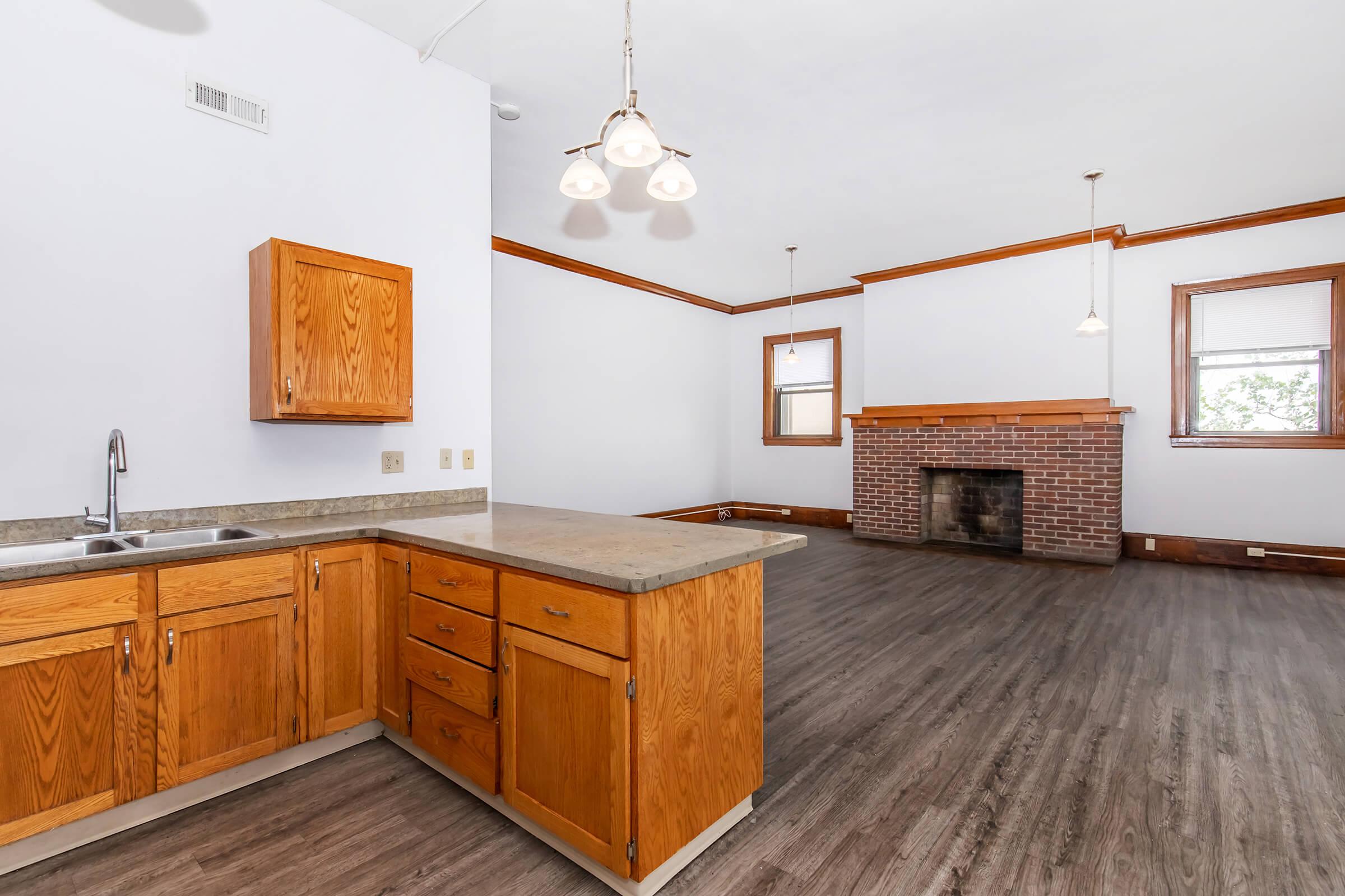 A spacious living area featuring a kitchen with wooden cabinets and a countertop. The room has large windows allowing natural light, wooden flooring, and a brick fireplace flanked by decorative lights. The open layout connects the kitchen and living space, creating a warm, inviting atmosphere.