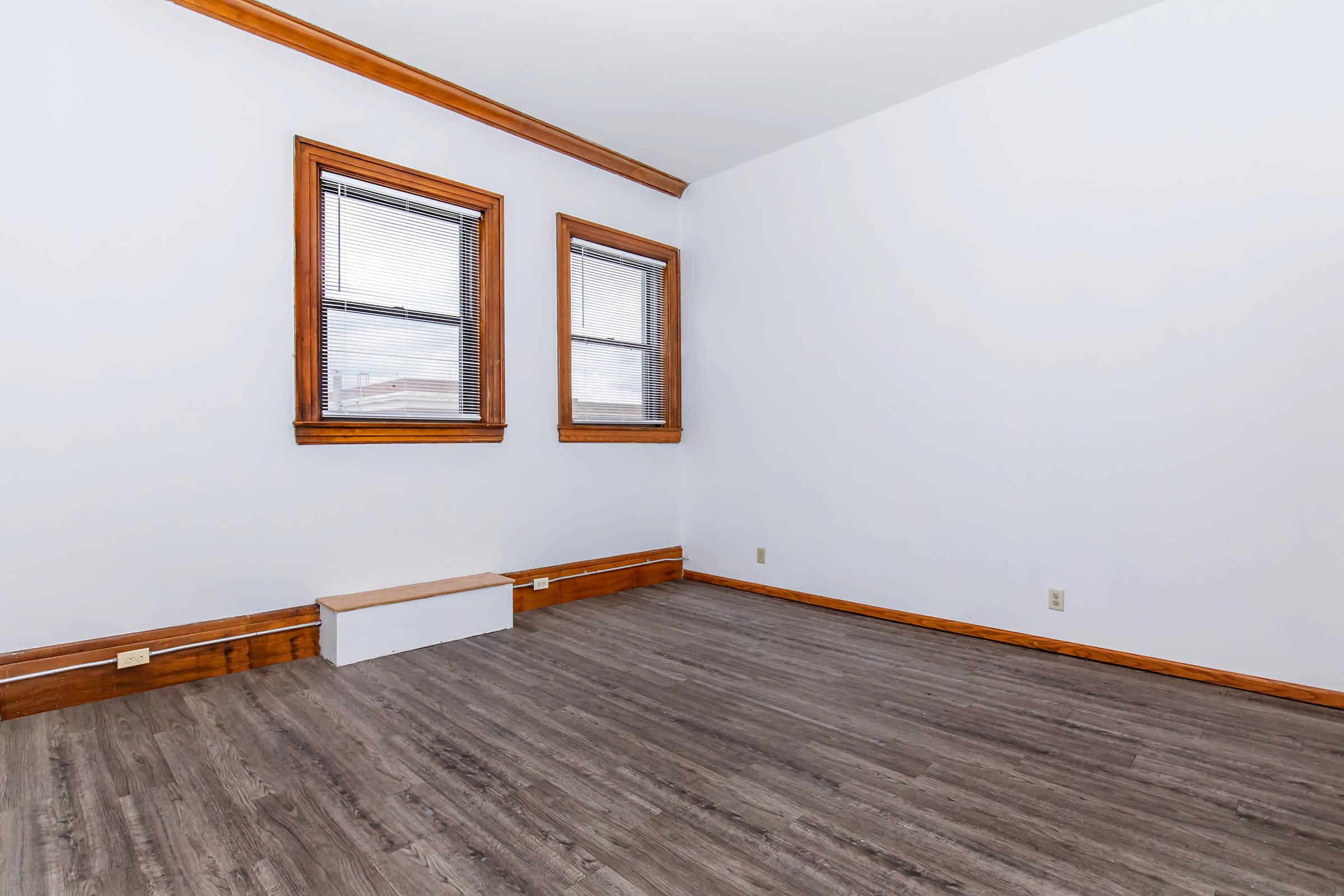 Empty room with white walls and wooden trim. Two windows with blinds on the left wall, and a small wooden ledge beneath them. The floor is covered with a light-colored laminate, giving a clean and open appearance. The space is well-lit and airy, suitable for various uses.