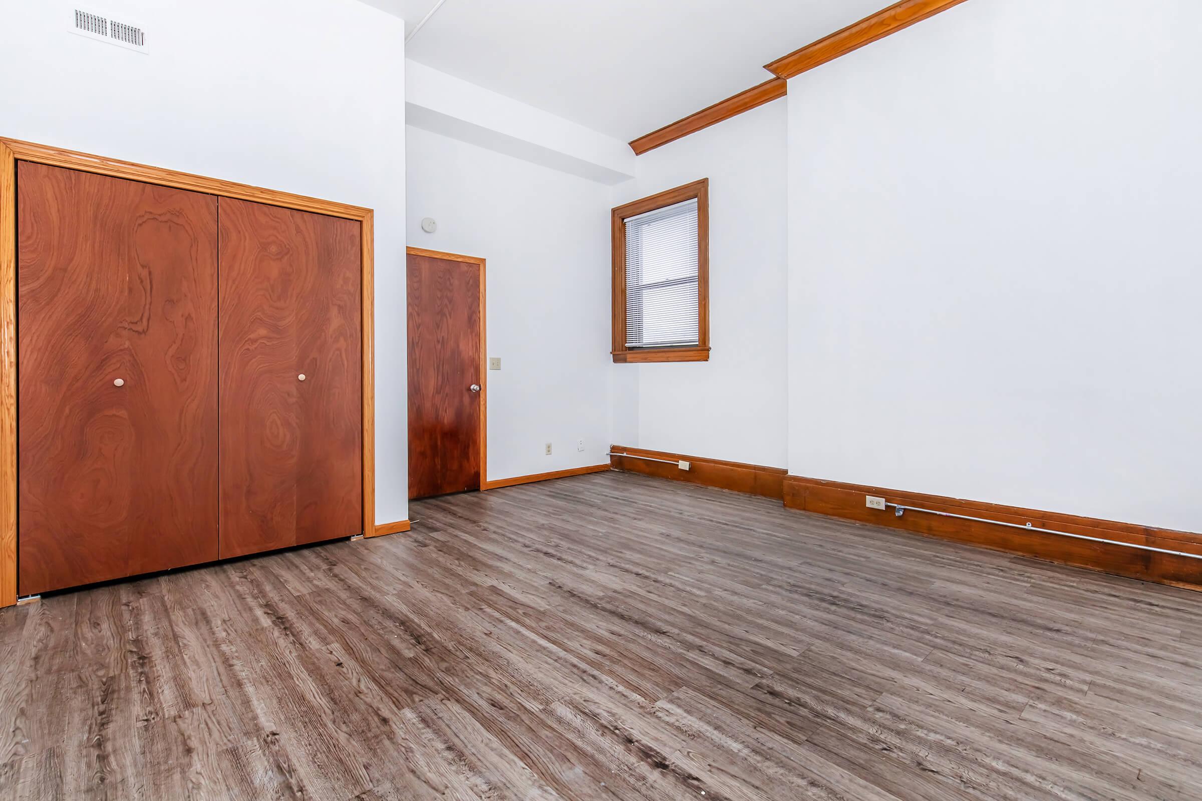 Interior of a room featuring hardwood floors and wooden trim. Two wooden doors are visible on the left side, with a window on the right wall allowing natural light. The walls are painted white, creating a bright and open atmosphere.