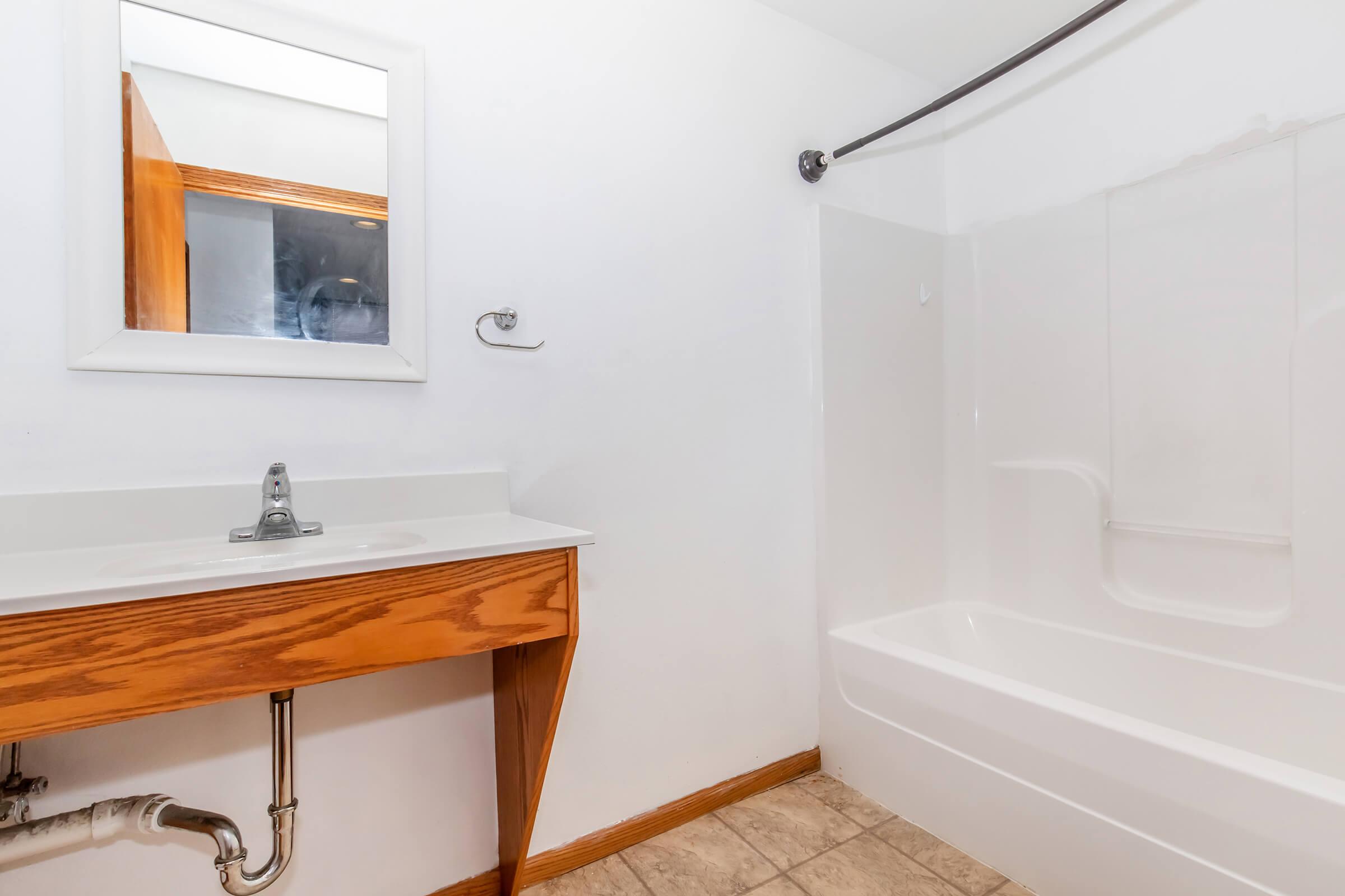 A small bathroom featuring a white bathtub, a wooden vanity with a sink, and a wall mirror. The walls are painted white, and the floor is tiled. A showerhead is installed above the tub, and there's a towel rack on the wall. The overall design is simple and functional.