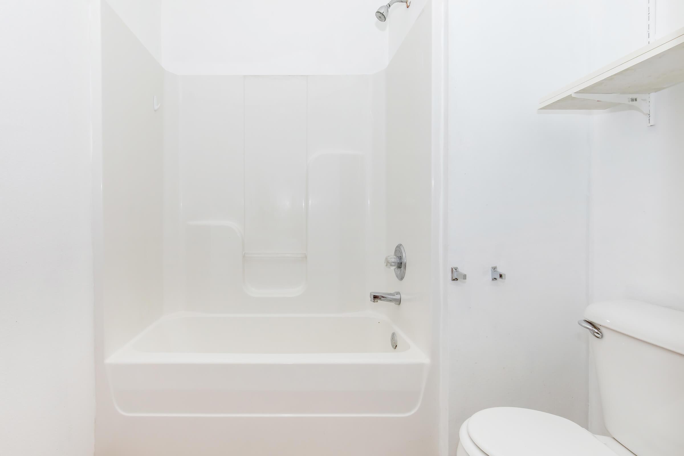A clean, modern bathroom featuring a white bathtub with a shower, a toilet, and a small shelf. The walls are plain white, creating a bright and minimalistic look.