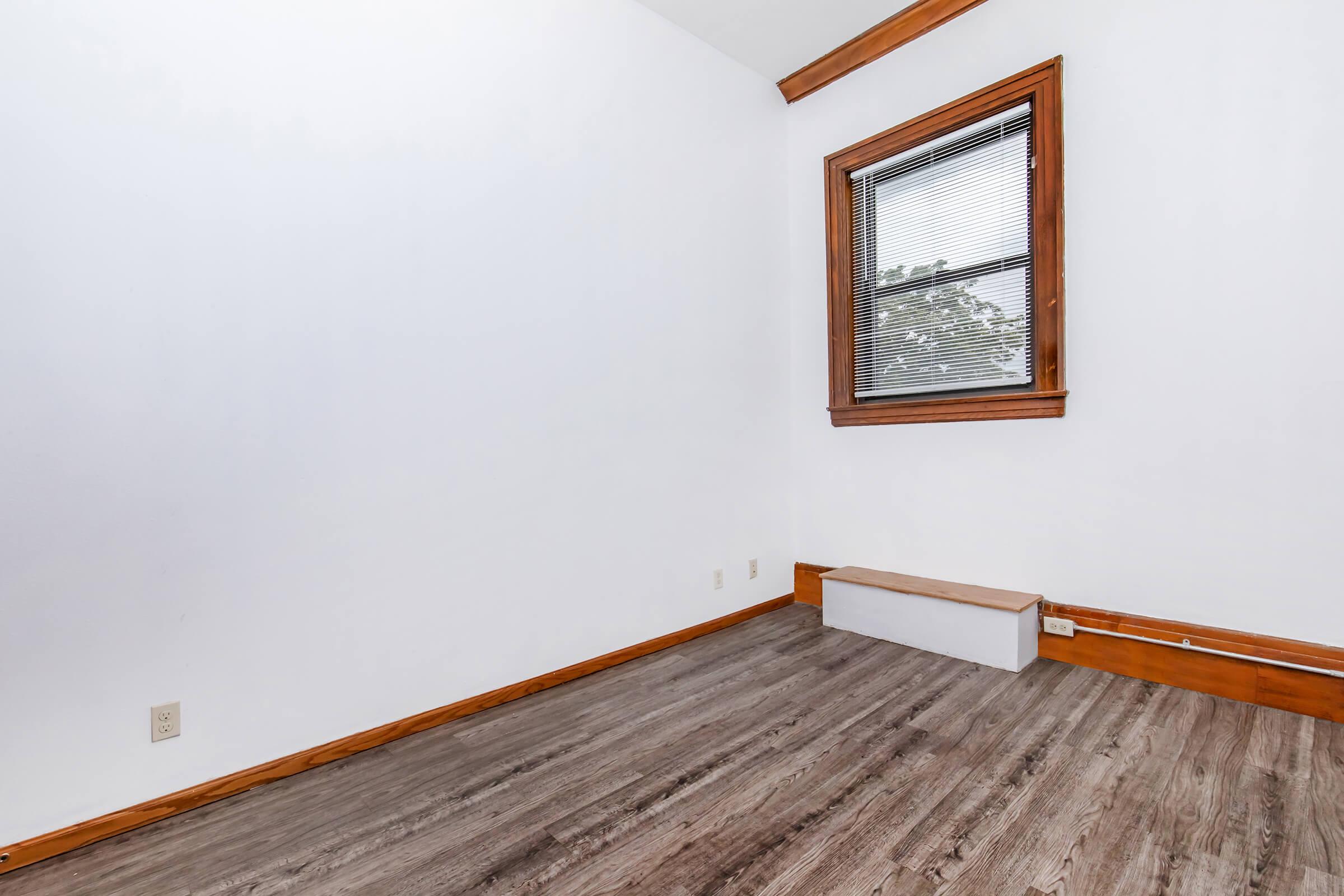 Empty room with white walls and wood trim, featuring a window with blinds and a small ledge below. The floor has a wood-like finish, and there are no furnishings in sight, creating a spacious and minimalistic atmosphere.