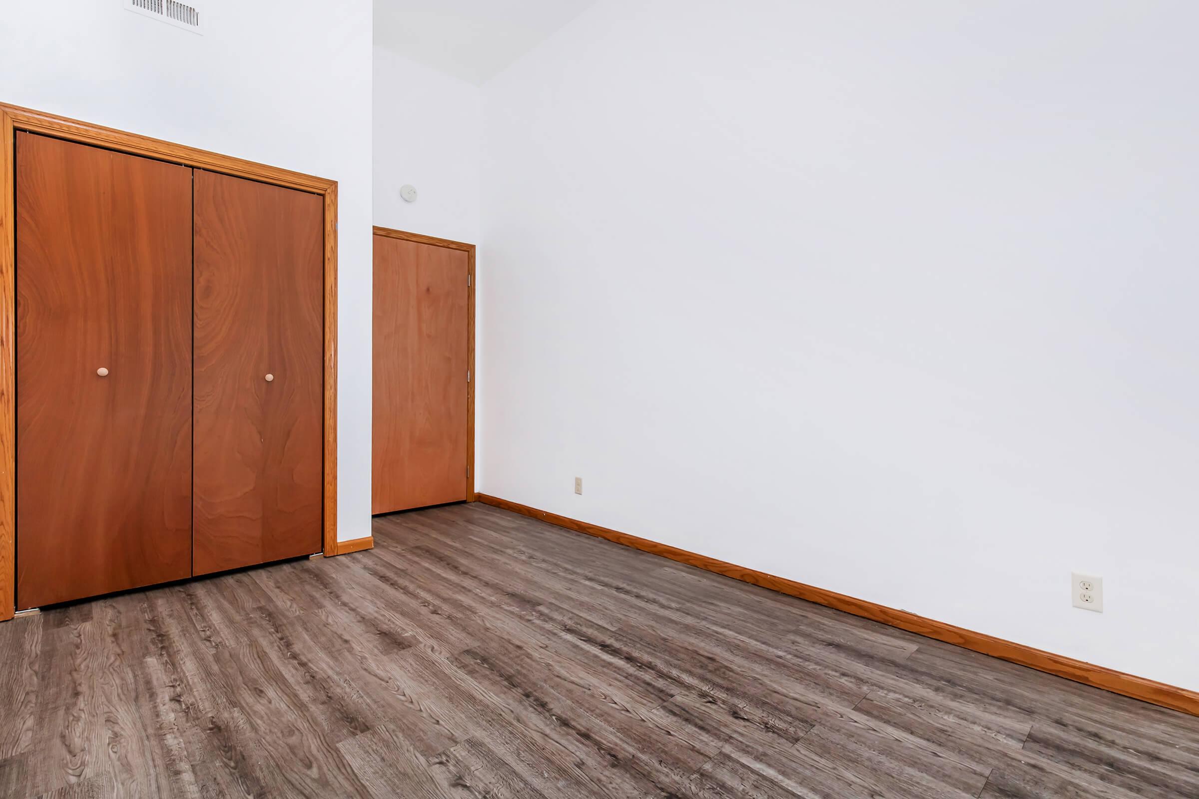 Empty room featuring light-colored walls and a wooden floor. There are two wooden doors on the left, one likely a closet and the other a regular door. The space is well-lit and has a minimalist design.
