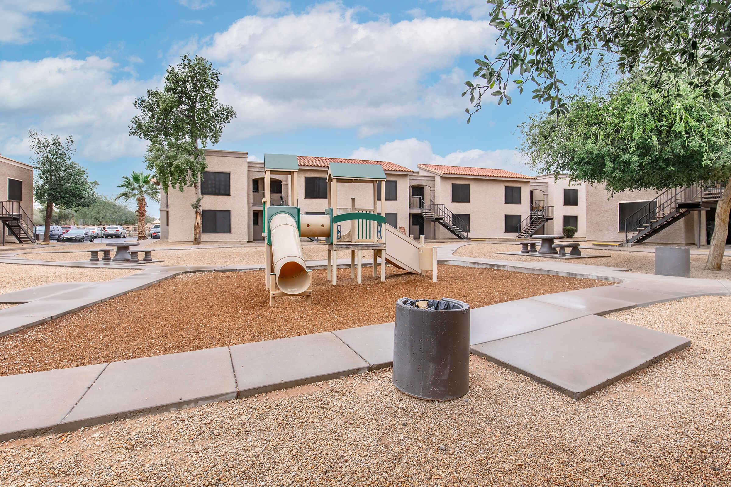 A playground featuring a slide and climbing structure, surrounded by gravel and grass. In the background, there are several apartment buildings and trees under a partly cloudy sky. A trash can is visible nearby on the paved walkway.