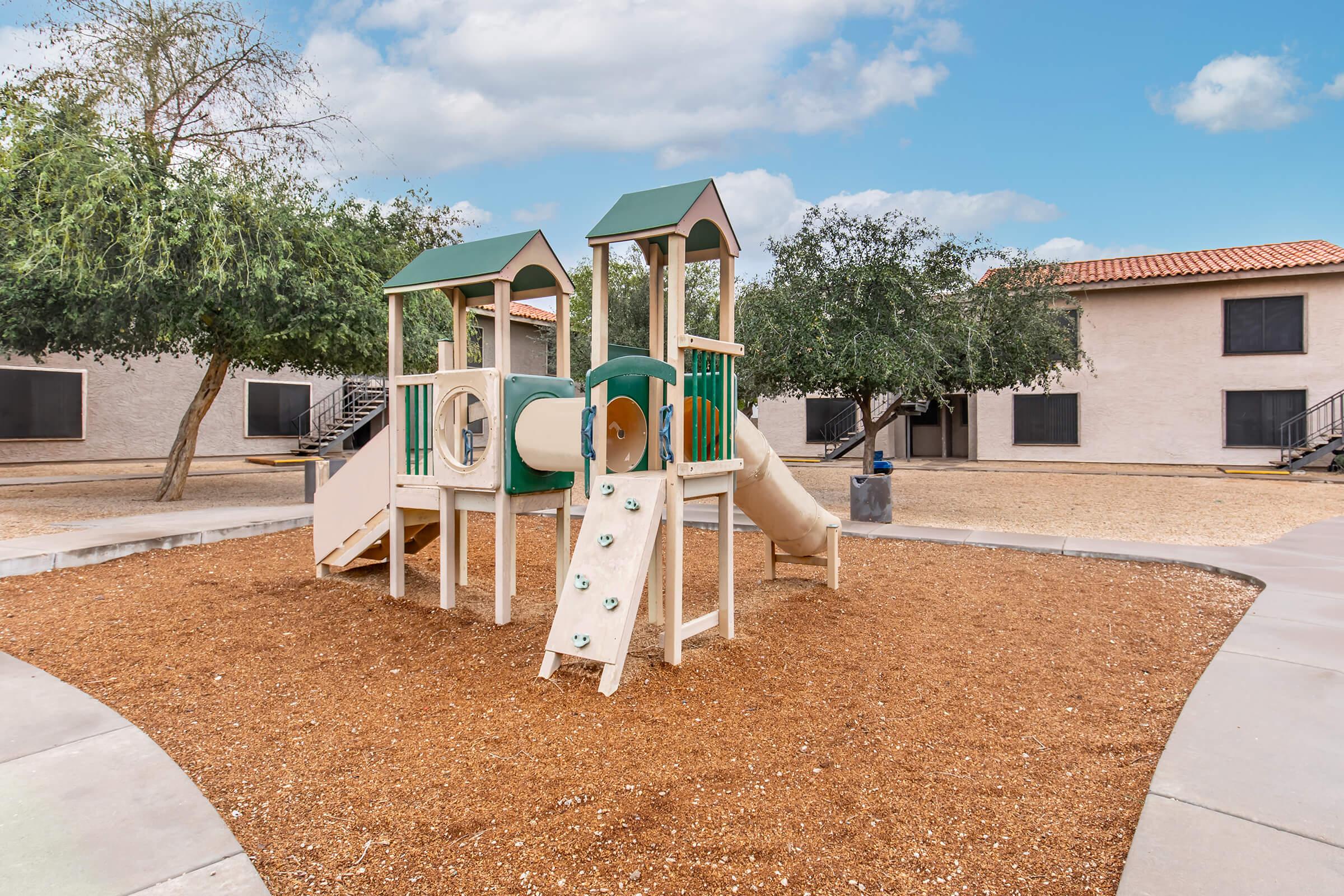 A children's playground featuring a small climbing structure with slides and climbing rocks, surrounded by a sandy area and enclosed by trees. In the background, there are residential buildings with windows and a clear blue sky dotted with clouds.
