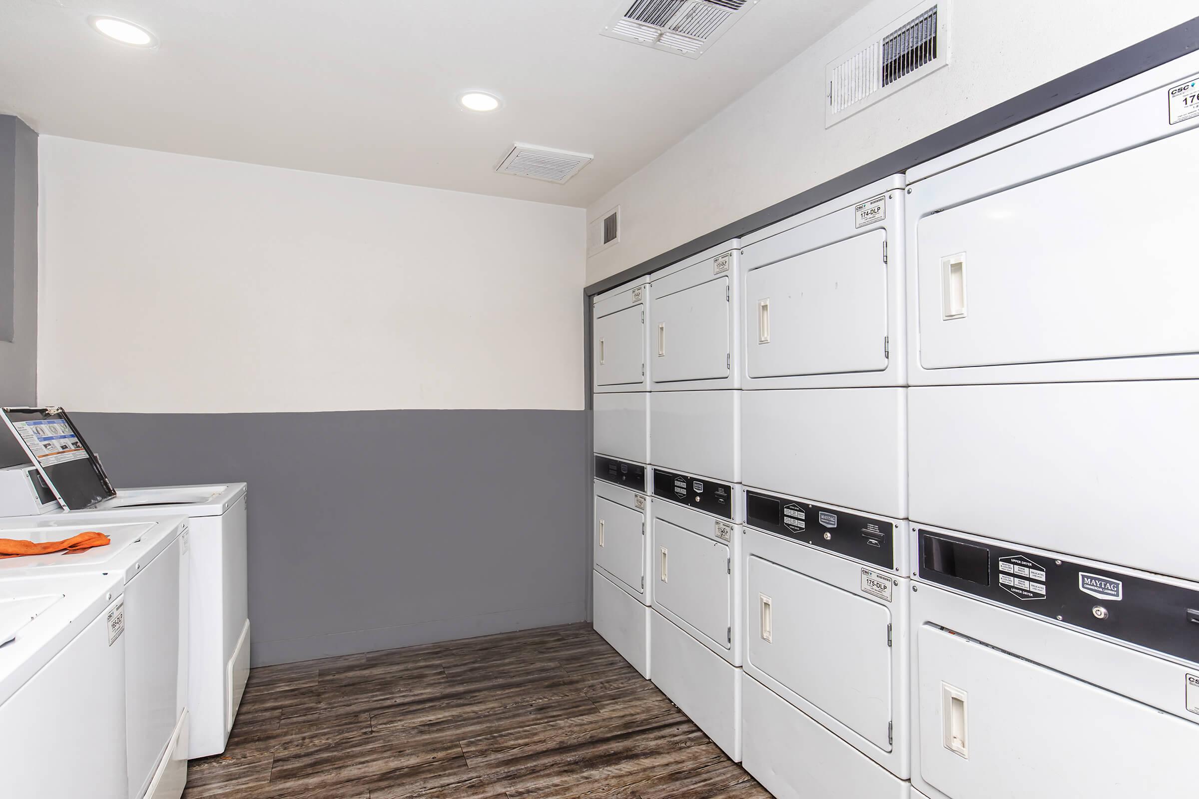 A clean and modern laundry room featuring several white washing machines and dryers arranged neatly along the wall. The room has light-colored walls with a gray section, bright lighting, and a wooden floor, creating a welcoming atmosphere for users.