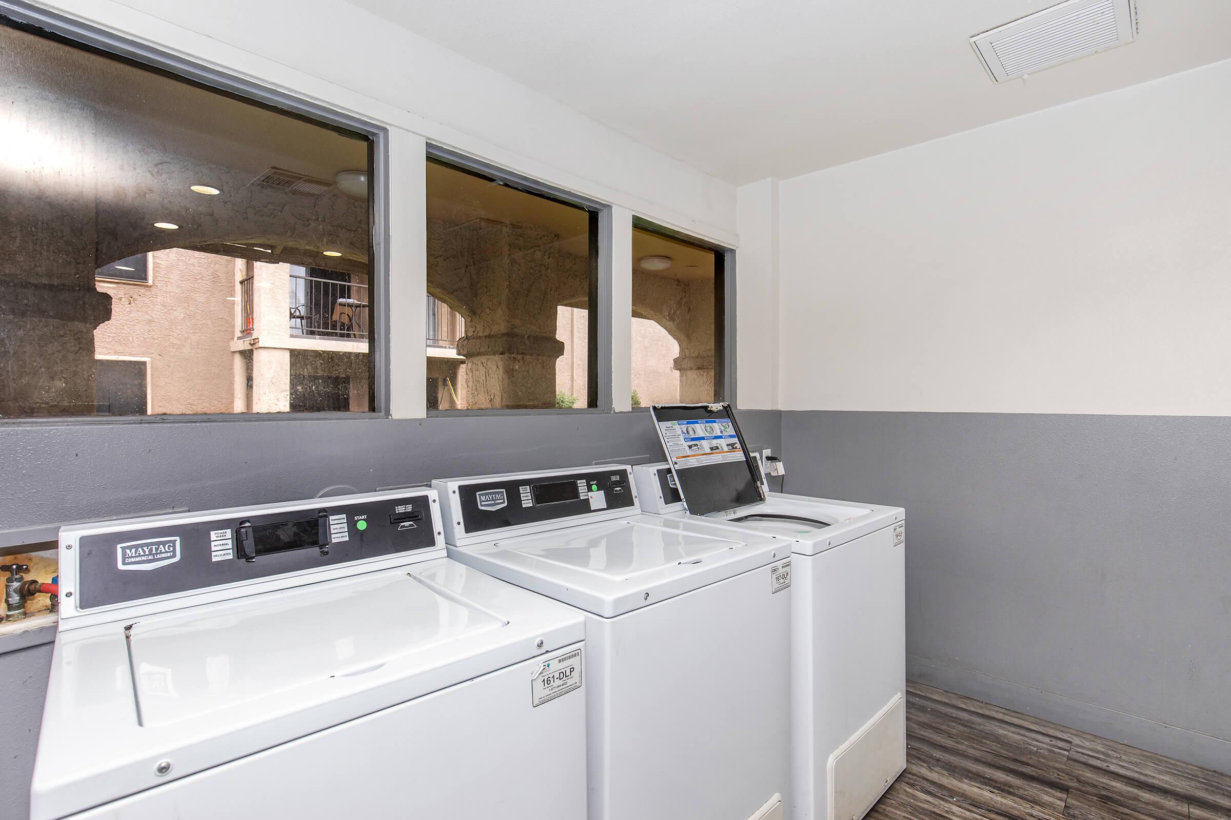 A laundry room featuring three white washing machines lined up against a wall. Large windows provide natural light, with a view of an outdoor area. The interior has neutral-colored walls with a gray bottom half and wooden-style flooring.