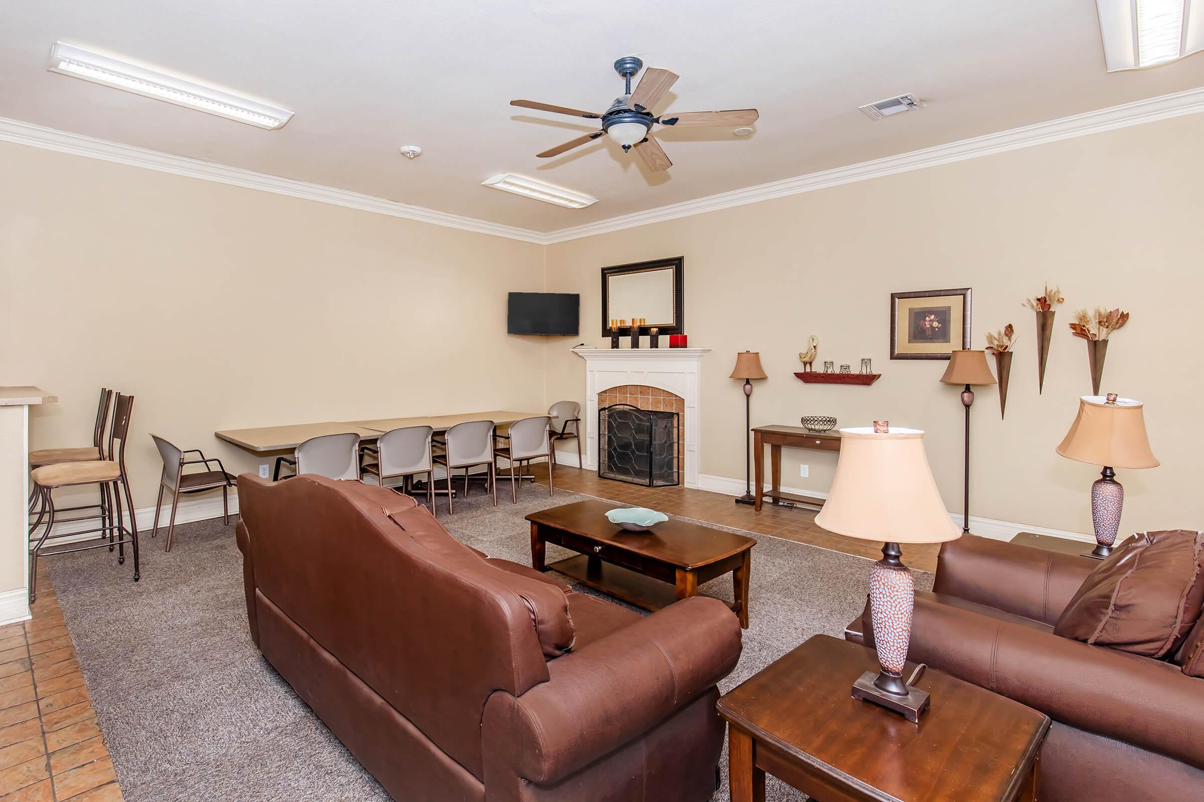 A cozy living area featuring two brown sofas and a wooden coffee table. There is a fireplace with a decorative mantel, a flat-screen TV mounted on the wall, and a dining table with chairs in the background. Soft lighting from table lamps adds warmth to the space. Neutral walls and carpet create a welcoming atmosphere.