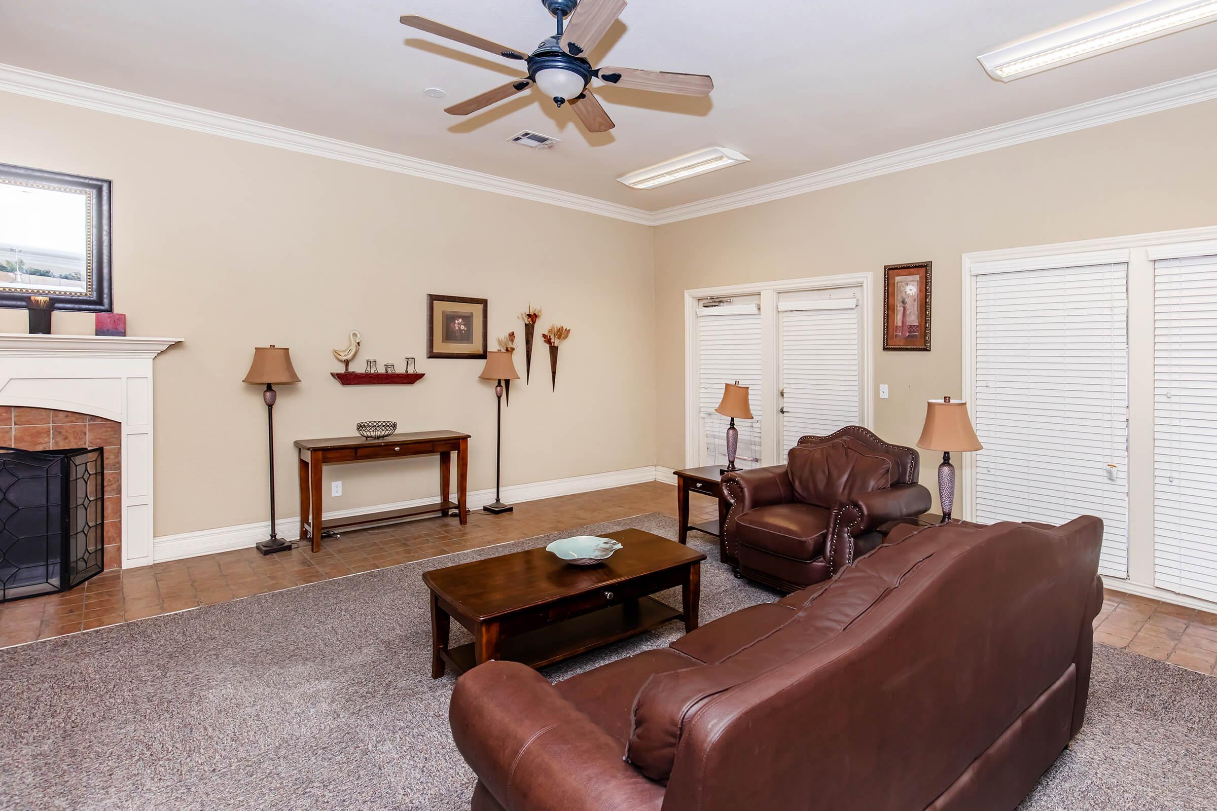 A cozy living room featuring two brown leather sofas, a wooden coffee table, and stylish lamps on either side. There's a fireplace with a decorative mantel, and light-colored walls enhance the space. Double doors with white shutters lead to another area, adding a touch of warmth and comfort.