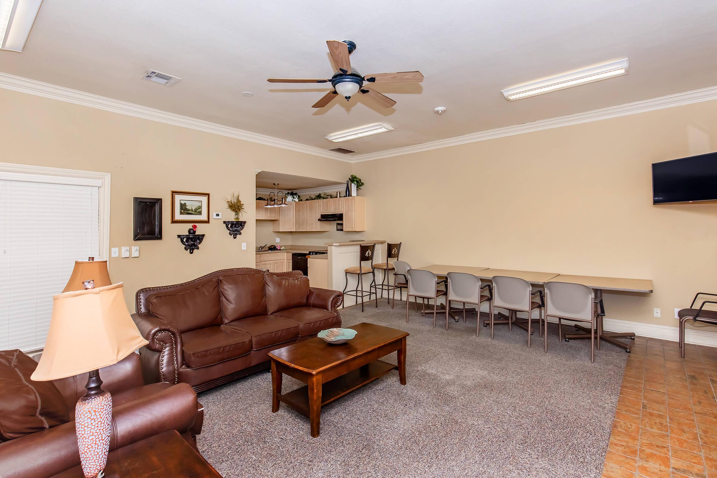A spacious room featuring a brown leather sofa and armchair, a wooden coffee table, and a ceiling fan. In the background, there is a kitchenette with light-colored cabinets and a countertop with bar seating. The walls are painted a warm beige, creating a cozy atmosphere.