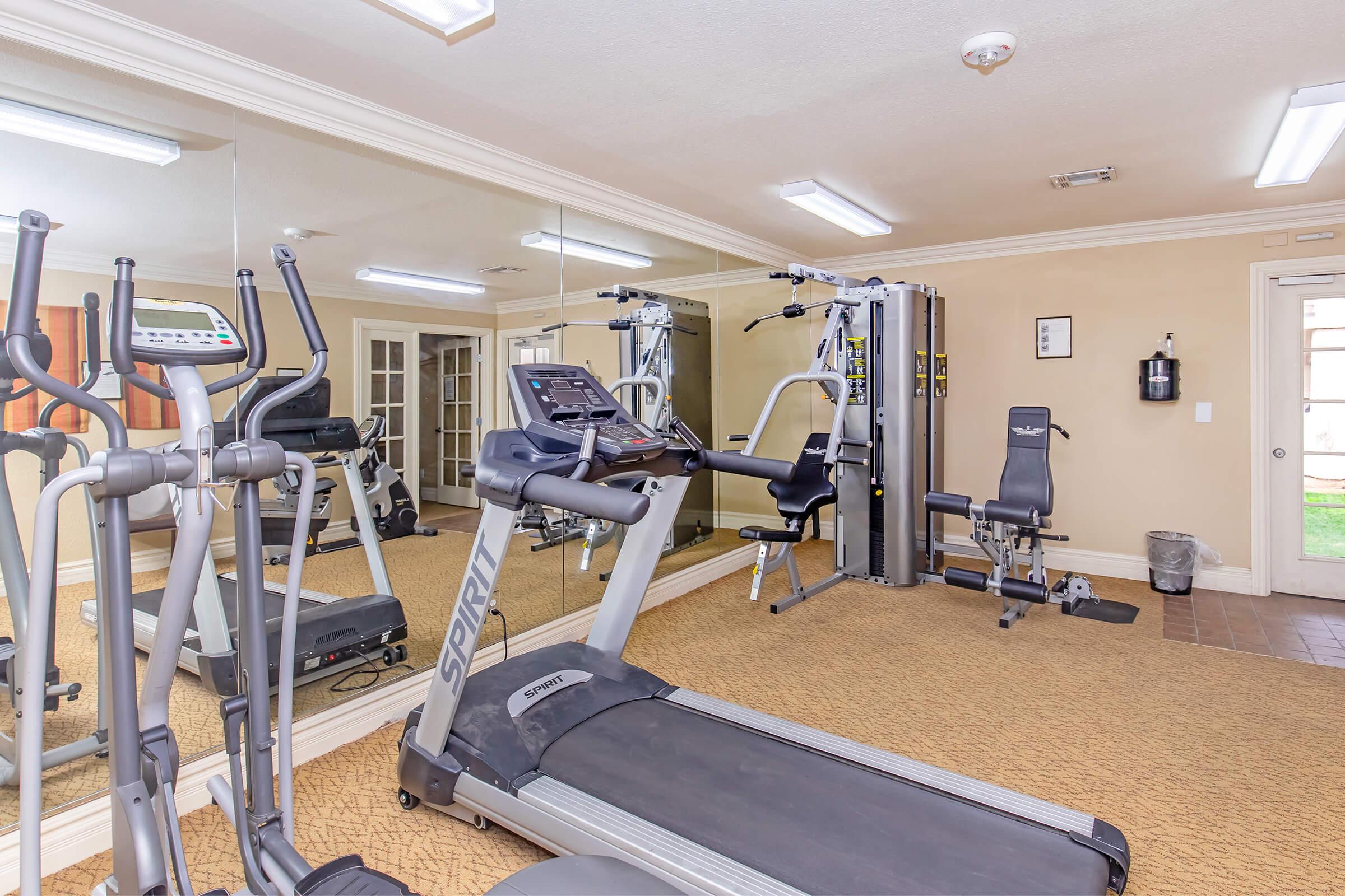 A spacious gym interior featuring various exercise equipment including a treadmill, a multi-gym machine, and exercise bikes. The area is well-lit with mirrors on one wall and a clean, welcoming atmosphere, perfect for workouts.