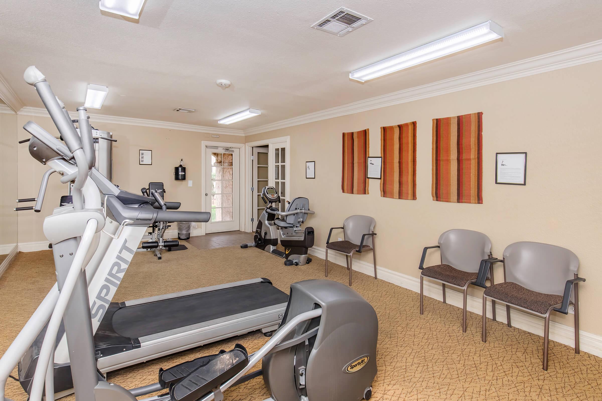 A fitness room featuring various exercise equipment, including a treadmill and an elliptical machine. There are chairs arranged along the walls, decorative artwork, and ample lighting, creating a welcoming space for workouts.