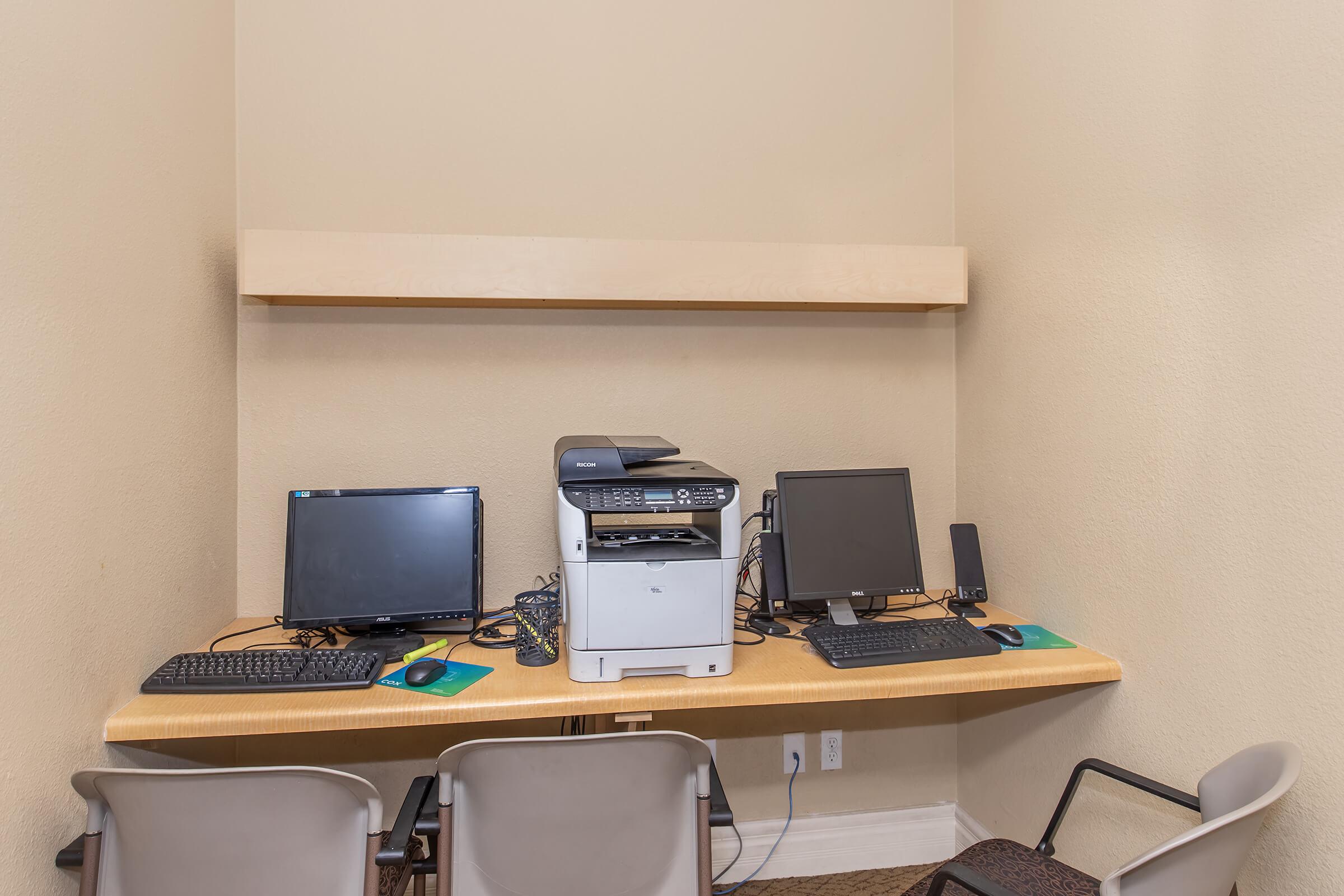 Two computer desks with black monitors and keyboards, a multifunction printer in the center, and a shelf above. There are three grey chairs in front of the desks, and the wall is painted a light color. The area appears organized and suitable for office or study use.