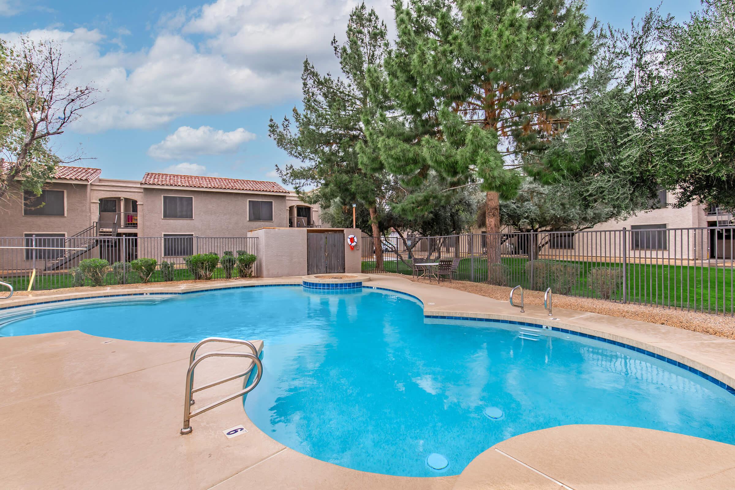 A serene swimming pool area with a curved edge, surrounded by well-maintained landscaping, trees, and a fence. Adjacent buildings are visible in the background, creating a relaxing atmosphere for residents. A lifebuoy is mounted nearby for safety.