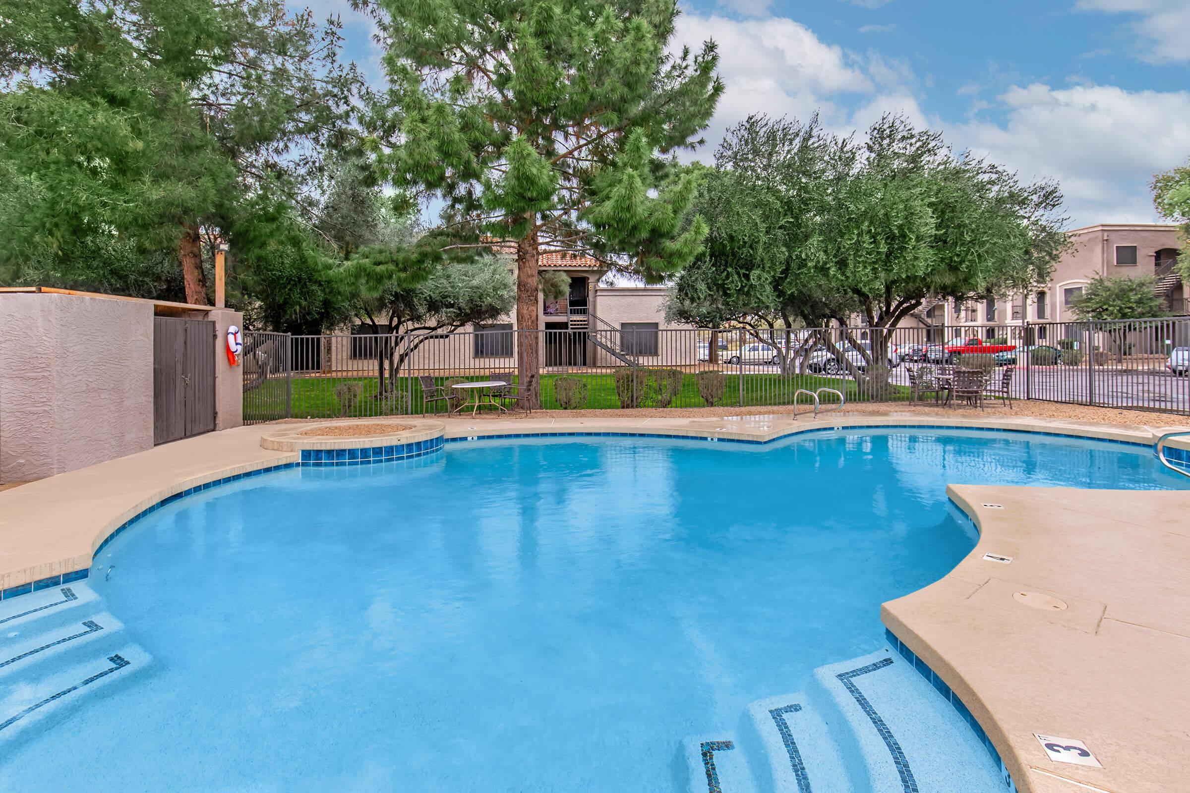 A swimming pool surrounded by greenery, featuring a curved design with steps leading into the water. In the background, there are trees and residential buildings. The sky is partly cloudy, creating a serene atmosphere.