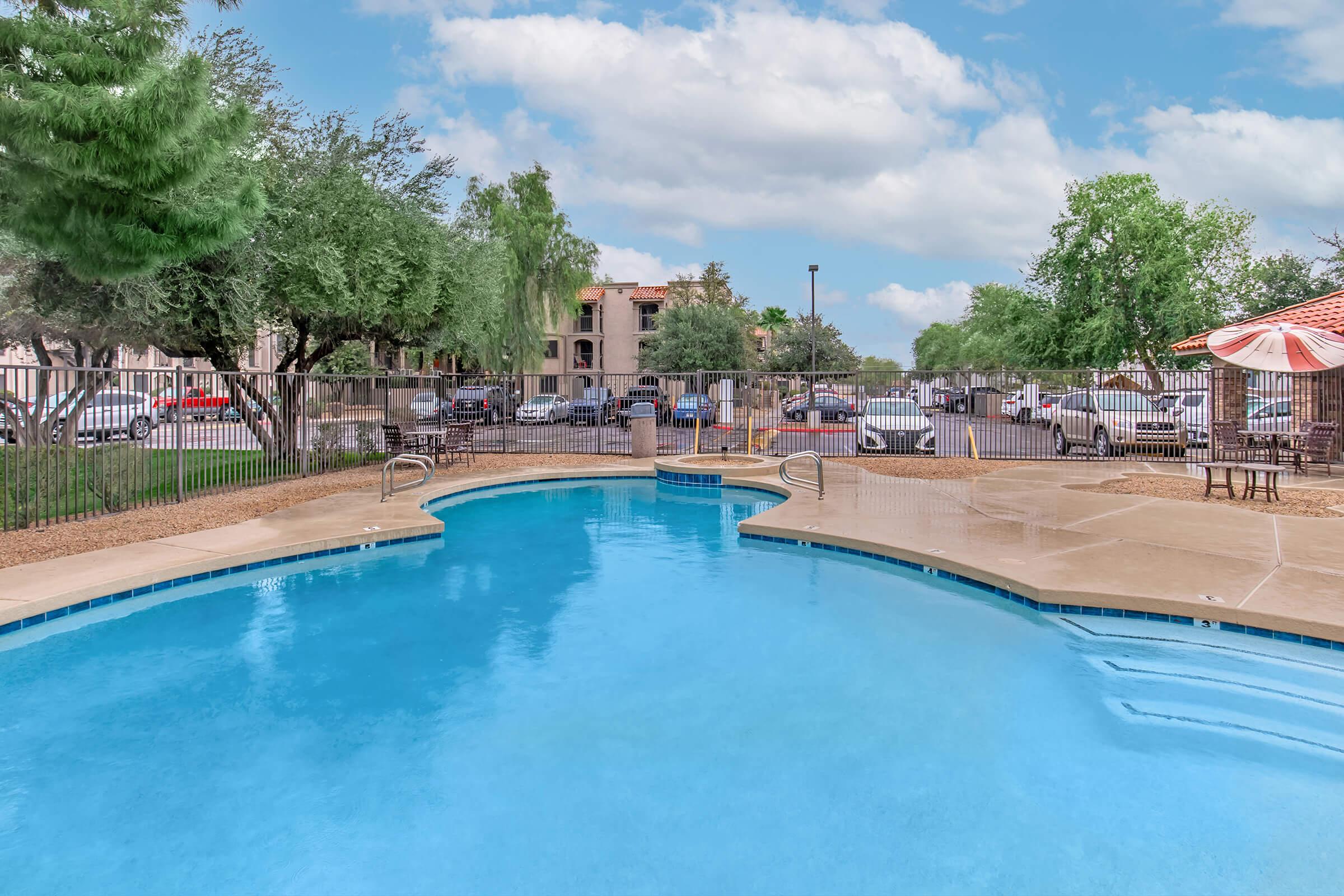 A clear blue swimming pool surrounded by a paved area, with lounge chairs and an umbrella. Lush green trees are visible nearby, and a fenced area separates the pool from a parking lot and apartment buildings in the background under a partly cloudy sky.
