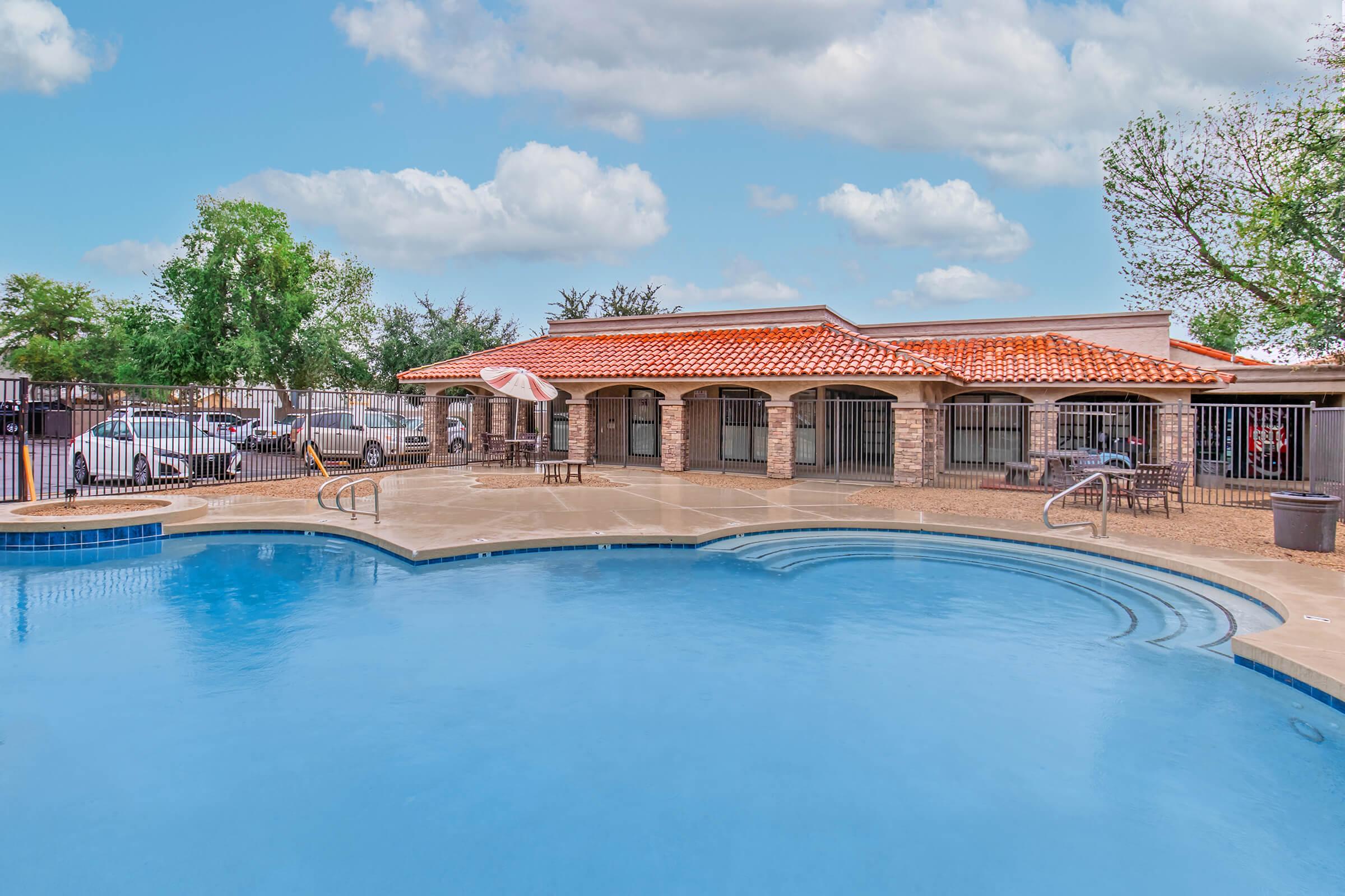A swimming pool with a curved edge and clear blue water, surrounded by a patio. There are a few lounge chairs, an umbrella for shade, and a brick building in the background with a red tiled roof. Trees and a fence are visible, along with parked cars nearby. The sky is partly cloudy.