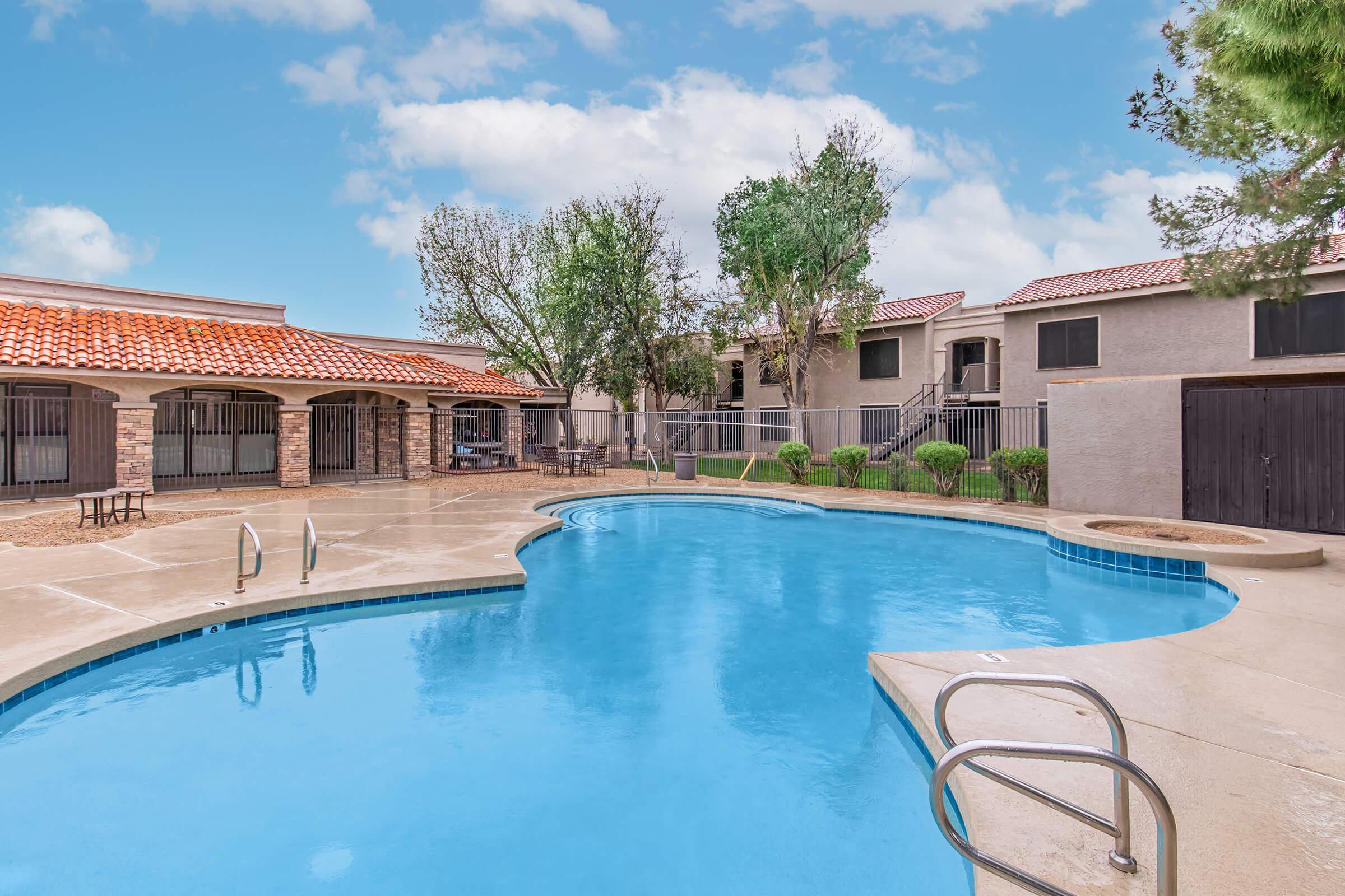 A clear blue swimming pool with a curved edge, surrounded by a concrete deck and lounge chairs. Lush green trees and shrubs are nearby, alongside a building with a red tile roof in the background. The sky is bright and partly cloudy, creating a serene atmosphere.