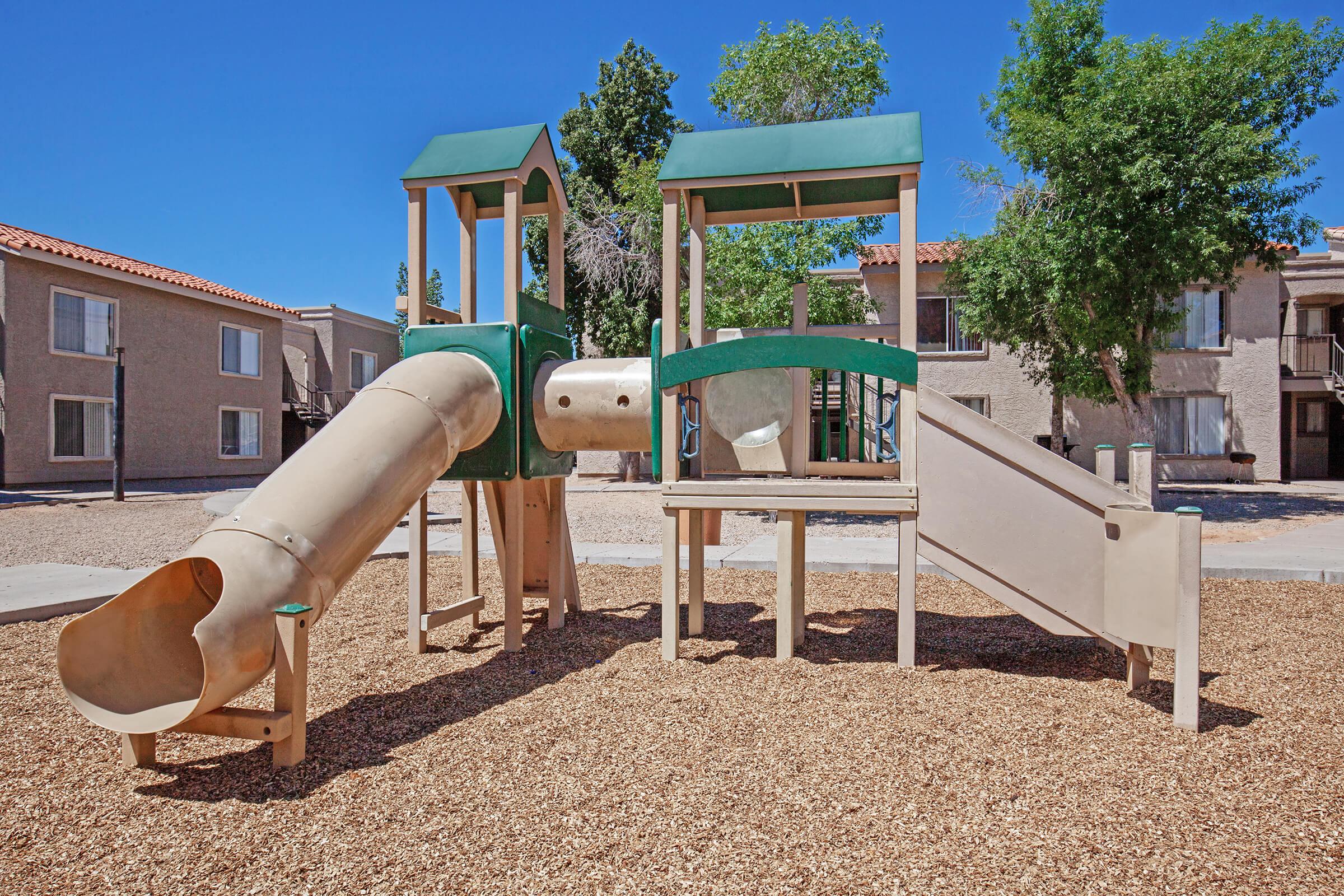 Playground structure featuring a beige slide, a small climbing area, and green roofs. The surrounding area has tan wood chips as ground cover, with residential buildings in the background. Clear blue sky and green trees are visible, indicating a sunny day.