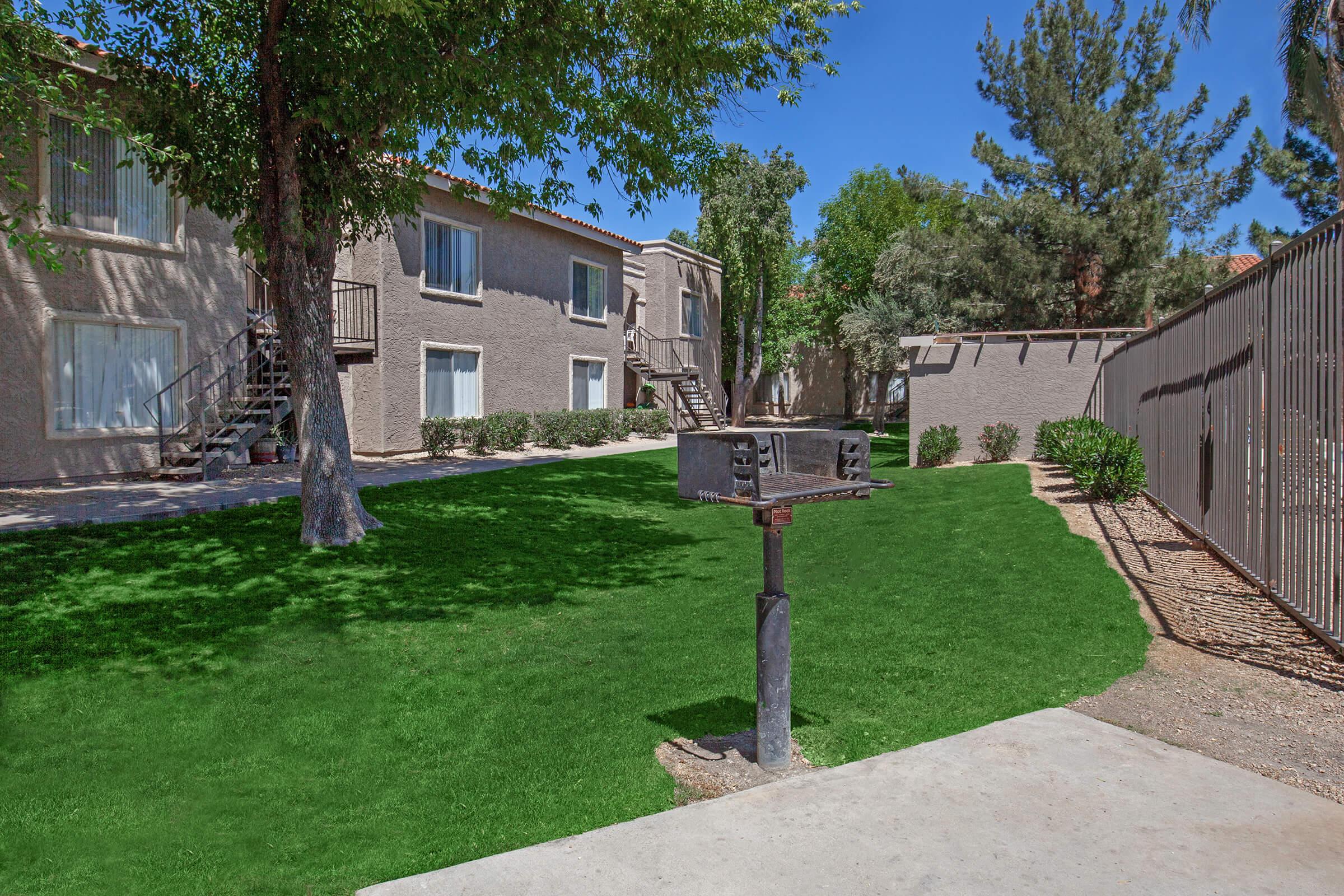 A sunny outdoor area featuring a well-maintained lawn with a grill near a walking path. In the background, there are two-story apartment buildings with balconies and greenery surrounding the space. Blue sky and trees provide a pleasant atmosphere for outdoor activities.