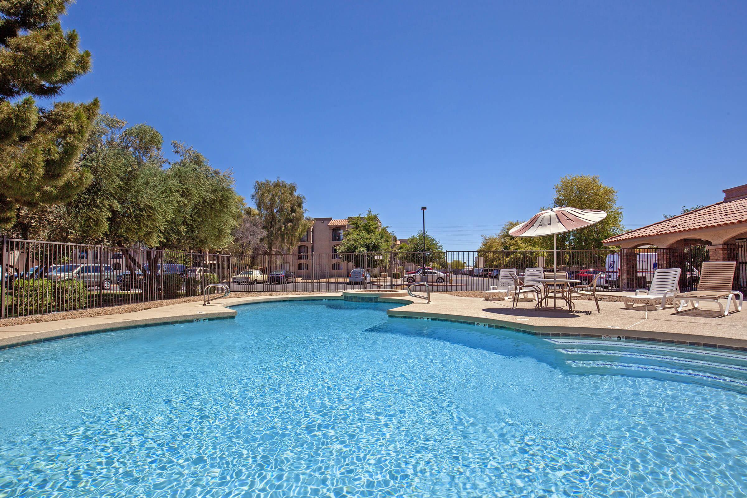 A clear blue swimming pool surrounded by lounge chairs, with trees and a few buildings in the background. An umbrella stands near the poolside, and the sky is bright and sunny.