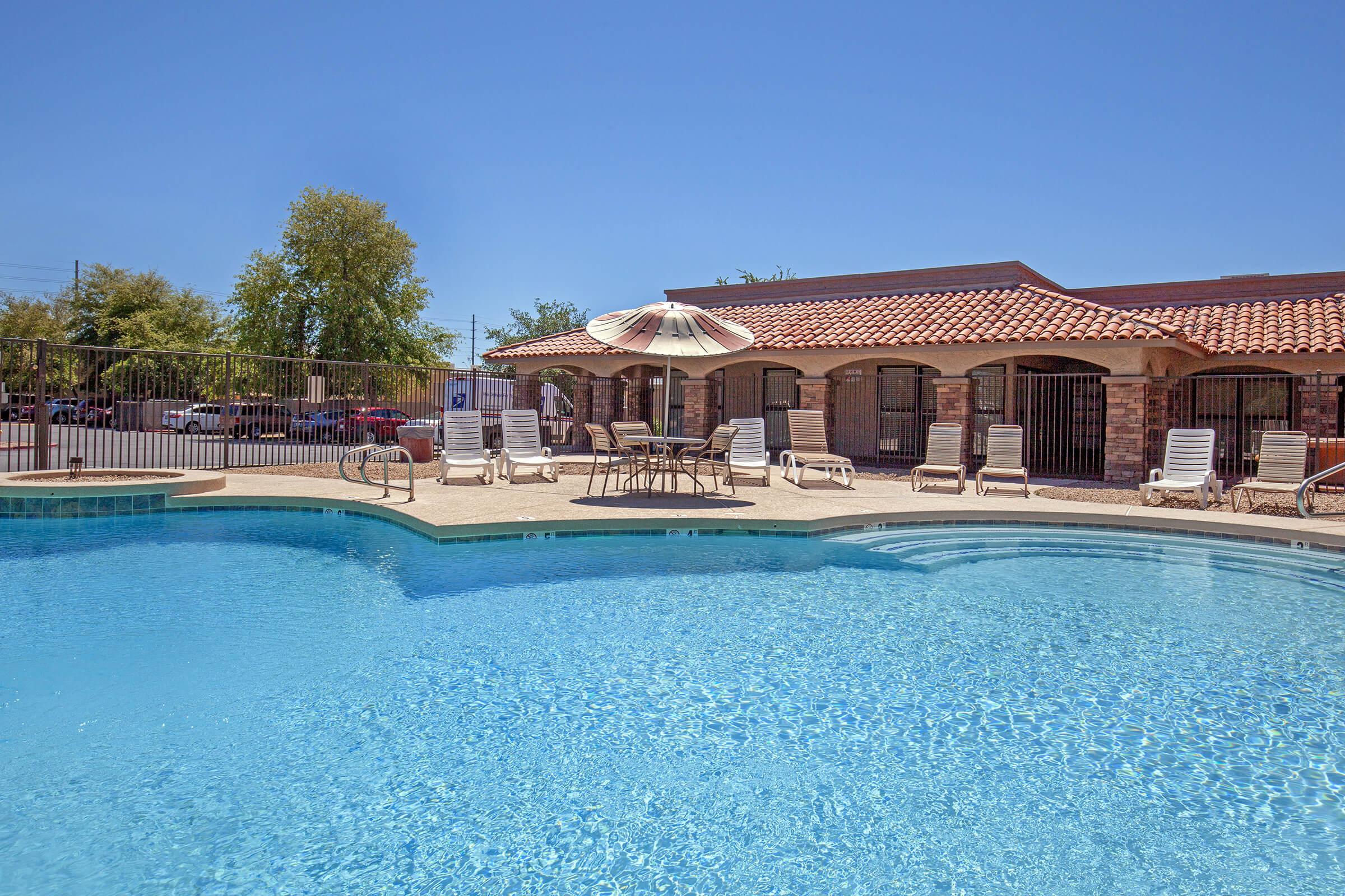 A sparkling blue pool surrounded by lounge chairs and an umbrella, with a sunny sky and a building in the background. The area is enclosed by a fence, providing a relaxing atmosphere perfect for leisure and enjoyment.