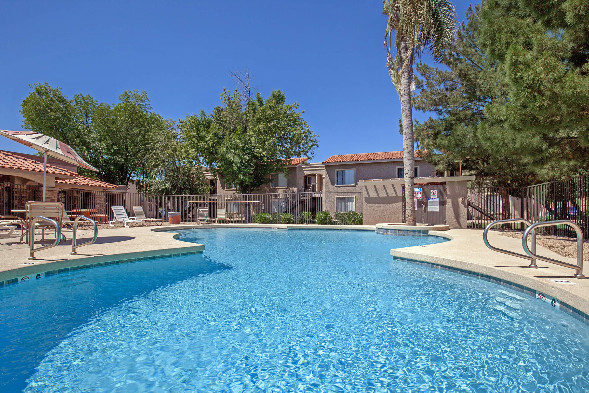 A clear swimming pool surrounded by palm trees and lounge chairs, with residential buildings in the background. The sky is blue and clear, creating a sunny and inviting atmosphere. The pool area is well-maintained, featuring a gentle curve and safety signage.