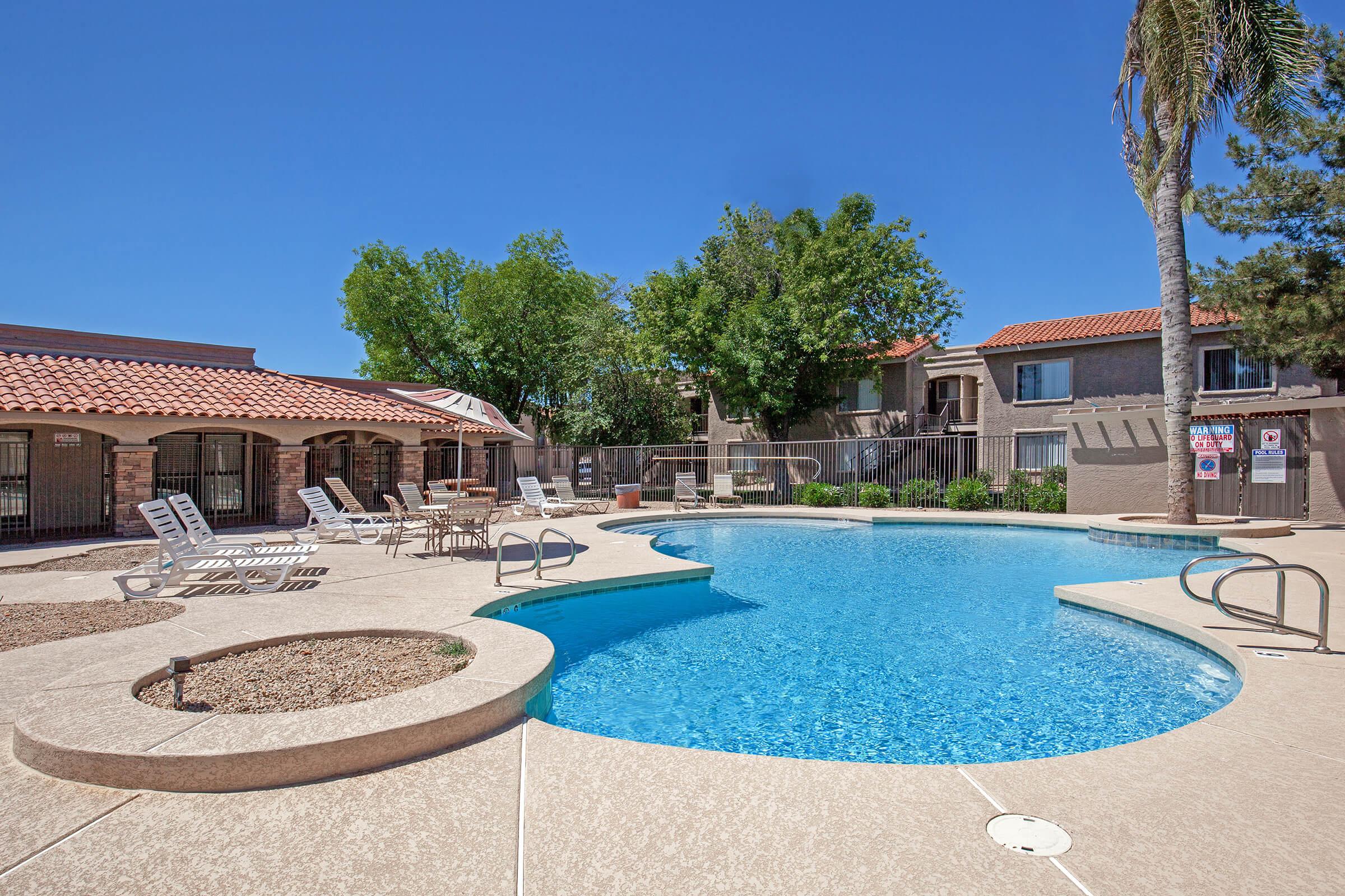 A bright outdoor pool area featuring a sparkling blue swimming pool surrounded by lounge chairs. Lush green trees provide shade, and there are nearby apartment buildings with terracotta roofs. Clear blue skies complete the inviting atmosphere of relaxation and leisure.