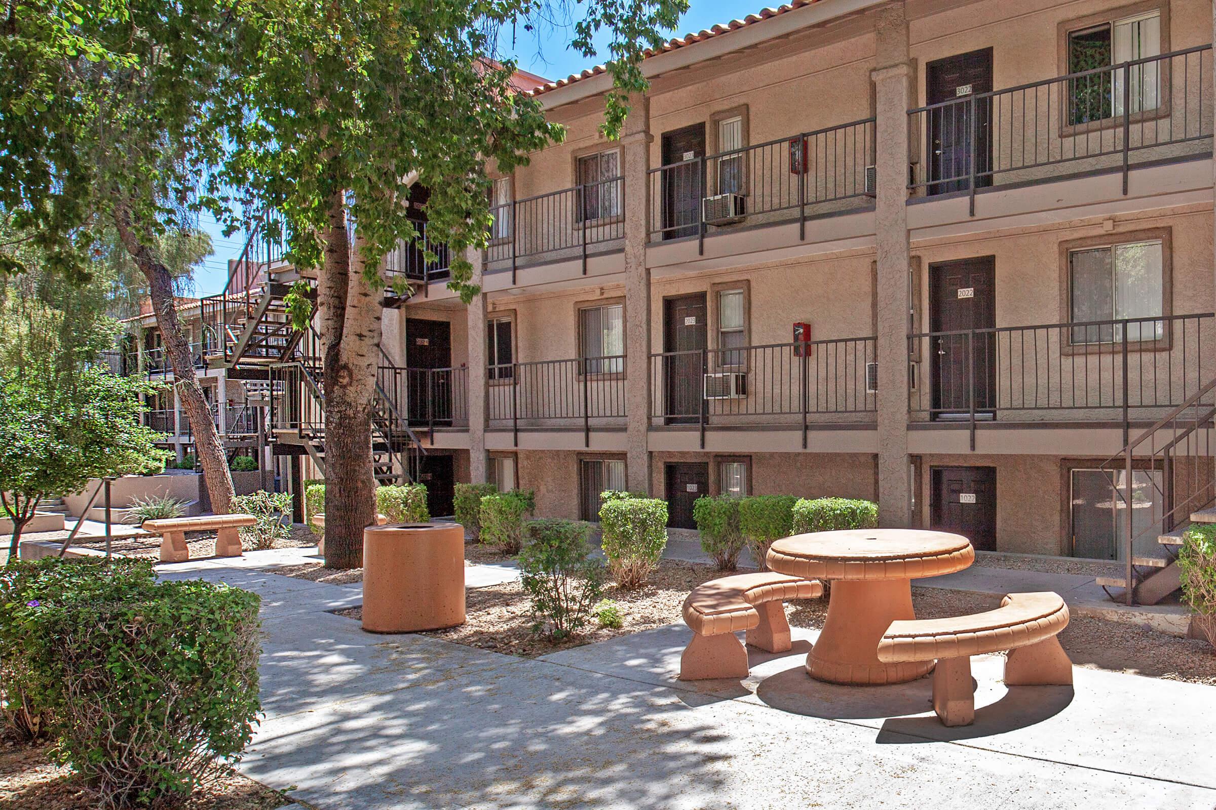 A view of a residential courtyard featuring multiple two-story apartment buildings. There are trees and neatly trimmed bushes surrounding a concrete pathway. In the center, a round picnic table with matching benches is located, providing a space for outdoor gatherings. Stairs lead to the upper levels of the buildings.