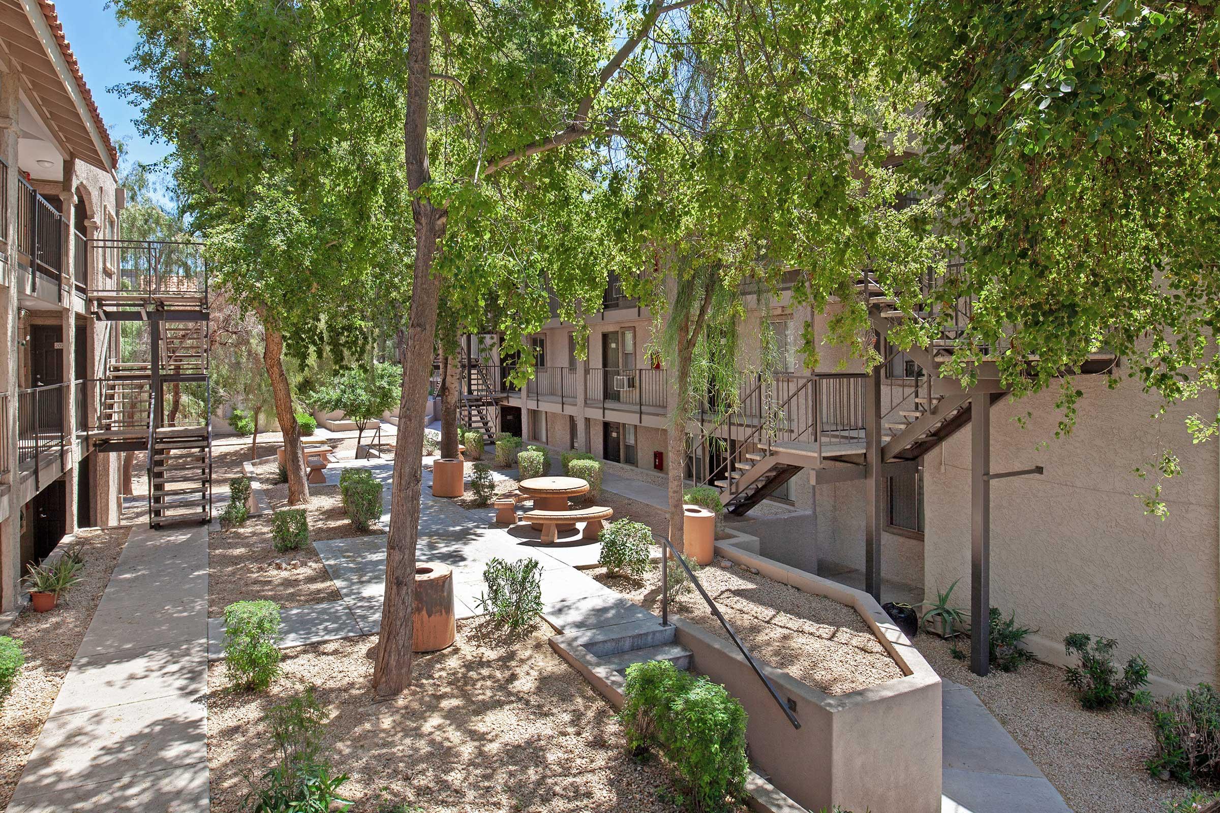 A landscaped courtyard featuring trees, shrubs, and gravel pathways. There are several buildings with staircases, outdoor seating areas, and planters, creating a tranquil atmosphere for residents.