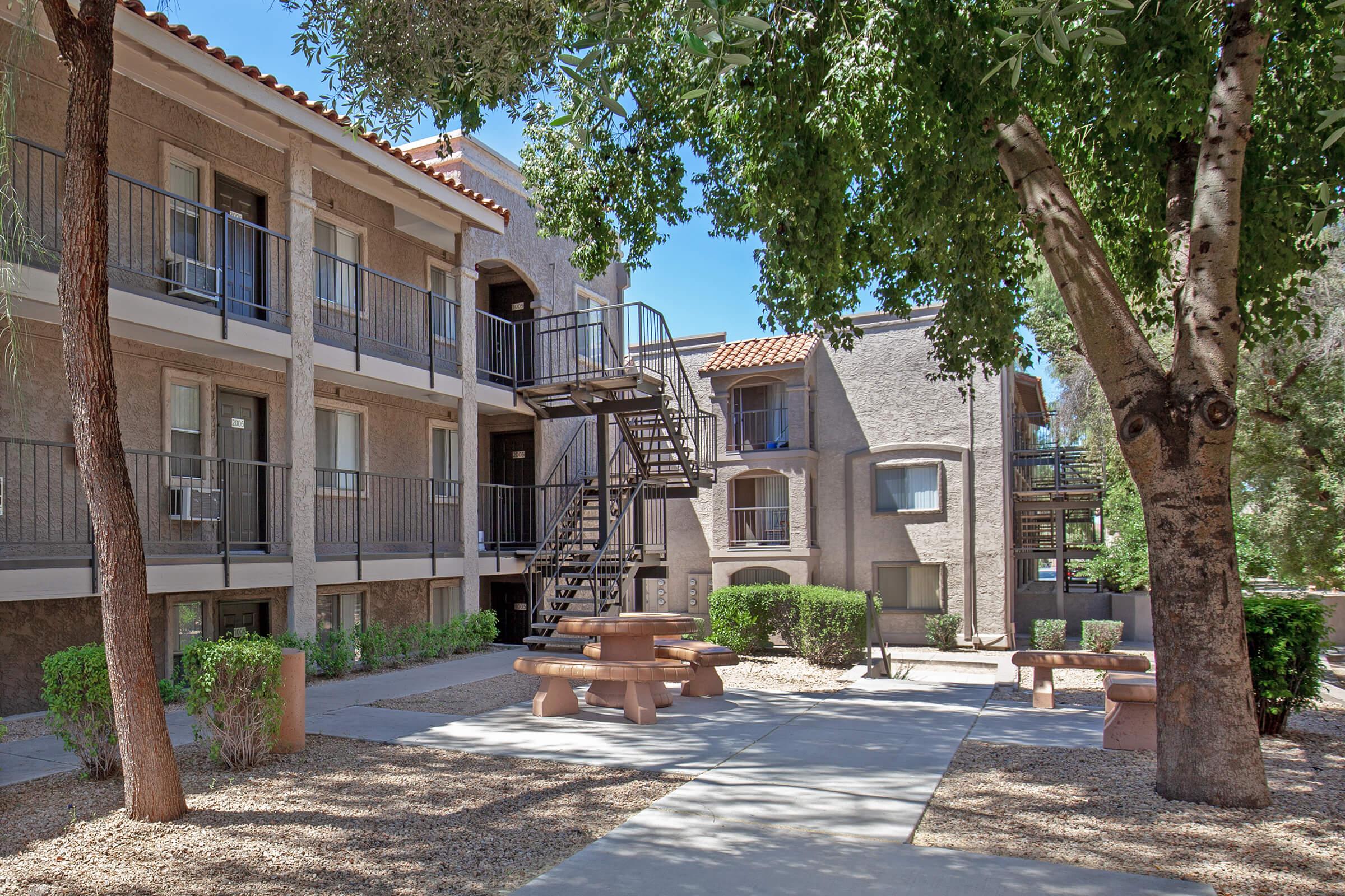A peaceful courtyard with trees and shrubs, featuring a gravel area with picnic tables. Surrounding the courtyard are two multi-story apartment buildings with balconies and a metal staircase visible. The scene is illuminated by bright sunlight, creating a welcoming atmosphere.