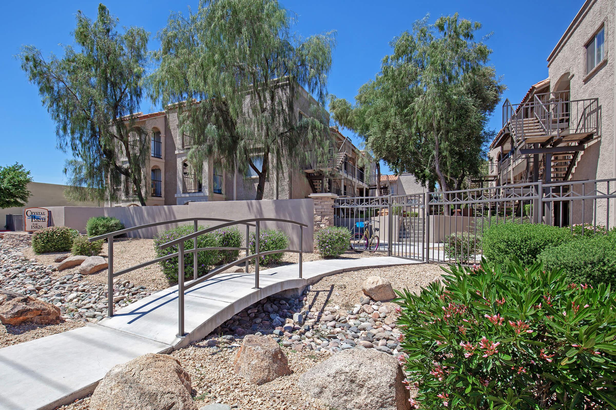 A serene outdoor scene featuring a walkway bridge leading through landscaped rocks and shrubs. In the background, there are multi-story apartment buildings partially obscured by trees, with a hint of outdoor staircases visible. Clear blue sky adds to the peaceful atmosphere.