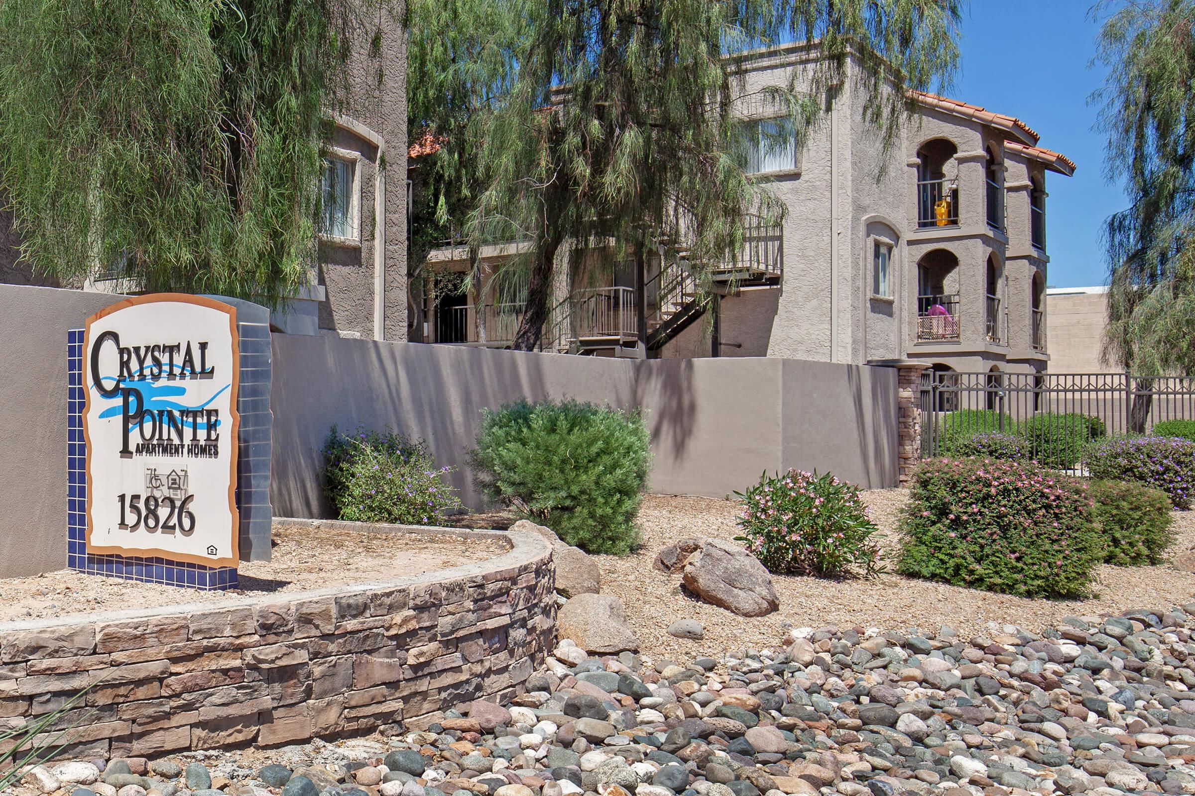 Exterior view of Crystal Pointe Apartments, featuring a decorative sign with the address 15826. The scene includes landscaped areas with bushes and a rocky path, alongside a multi-story building with balconies. Clear blue skies indicate a sunny day.