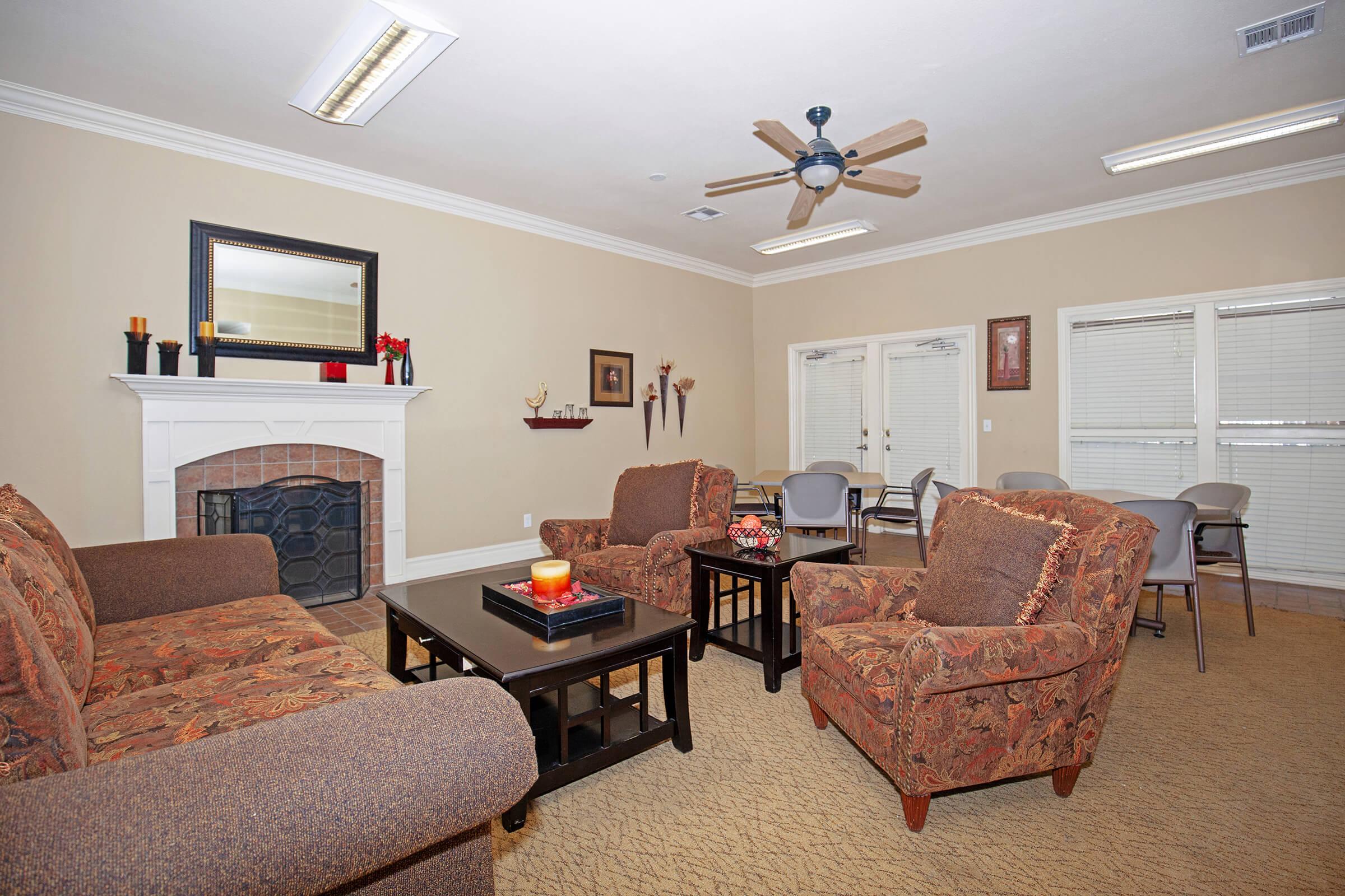 Cozy living room featuring two patterned sofas, a dark coffee table with a candle, and a decorative fireplace. There are two chairs and a dining area with four chairs in the background. The walls are painted a warm beige, and natural light flows in through two windows with blinds.