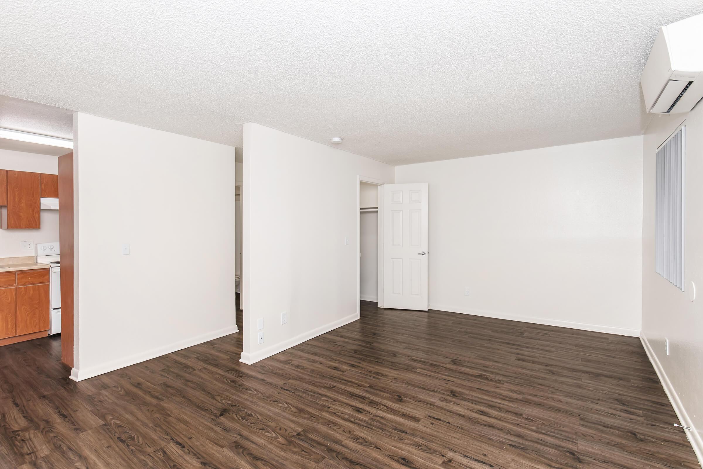 Empty room with light-colored walls and dark wood laminate flooring. A doorway leads to a closet on the right, while an open kitchen area is visible in the background. Natural light enters through a window, giving the space a bright and airy feel. Air conditioning unit mounted on the wall.