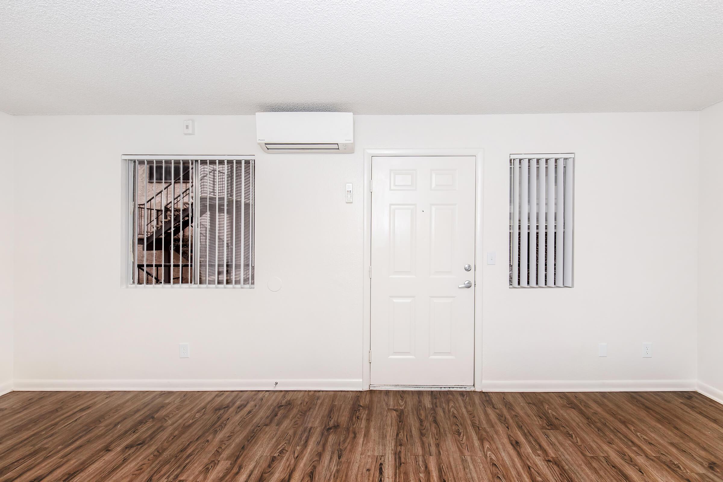 Empty room with light-colored walls and a wooden floor. A door is centered on the wall, flanked by two windows with metal bars. An air conditioning unit is mounted above the door. The space is minimally decorated and well-lit, offering a simple and clean aesthetic.