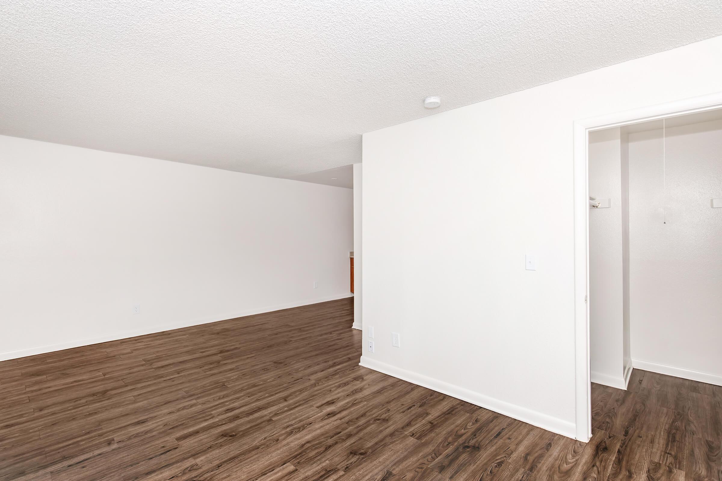 A spacious, empty room with light-colored walls and dark wood-style flooring. A doorway leads to another area, indicating potential for various setups. Natural light may enhance the space, offering versatility for furniture arrangement. The overall ambiance is clean and inviting.