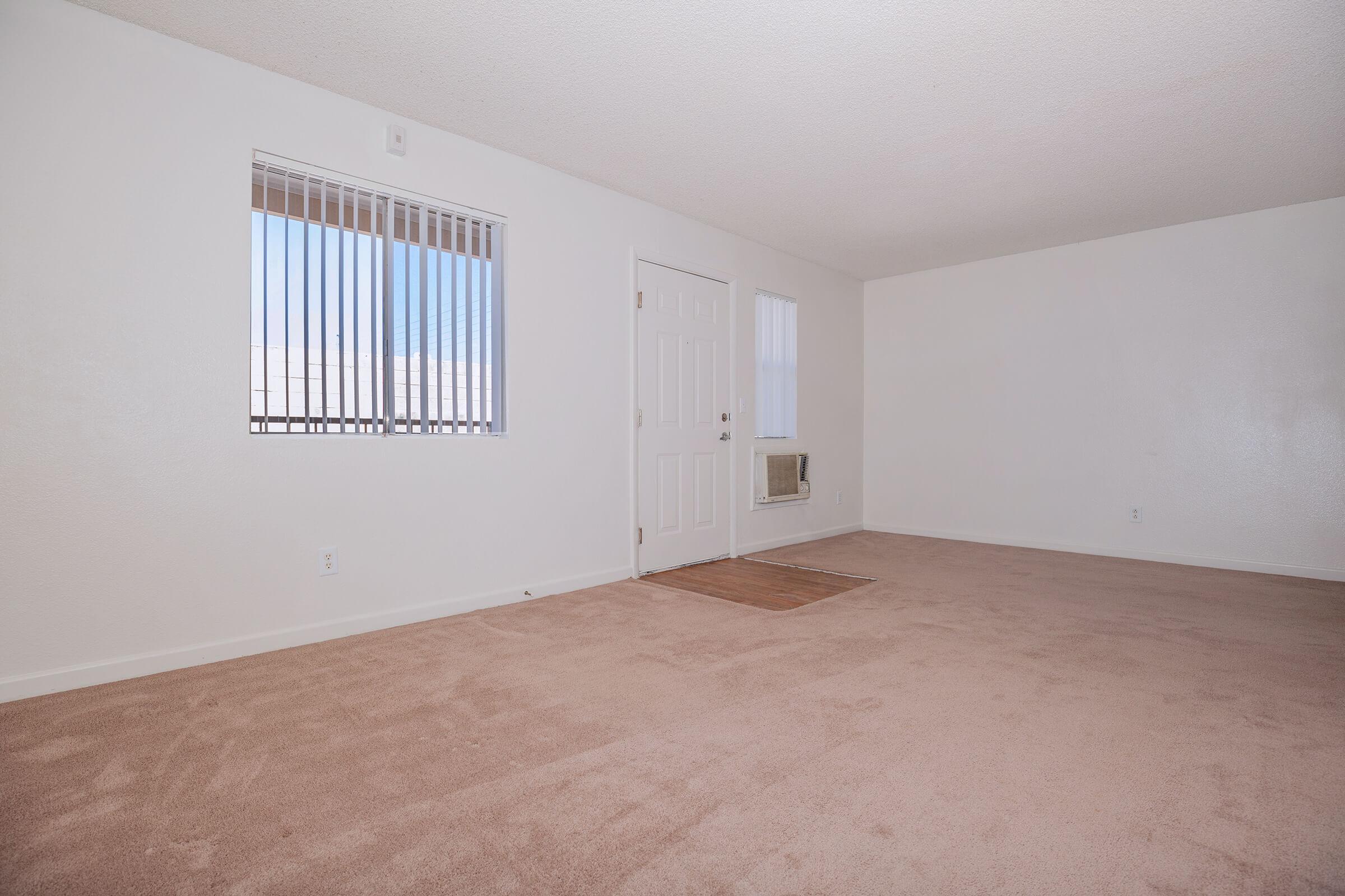 Empty room with beige carpet, white walls, and a window with vertical blinds. The door is located on the left, and there is an air conditioning unit mounted on the wall. Natural light enters through the window, creating a bright and spacious feel.