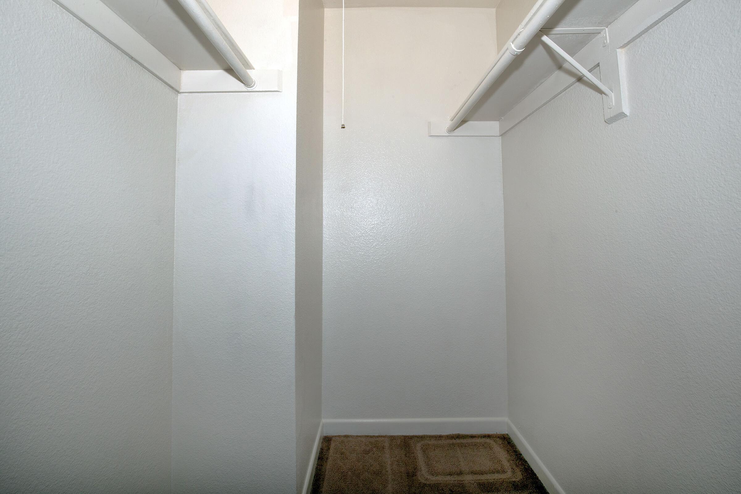 Empty closet with white walls and two open hanging rails. The floor is covered with a small beige mat. The space appears well-lit and clean, offering a minimalistic design.