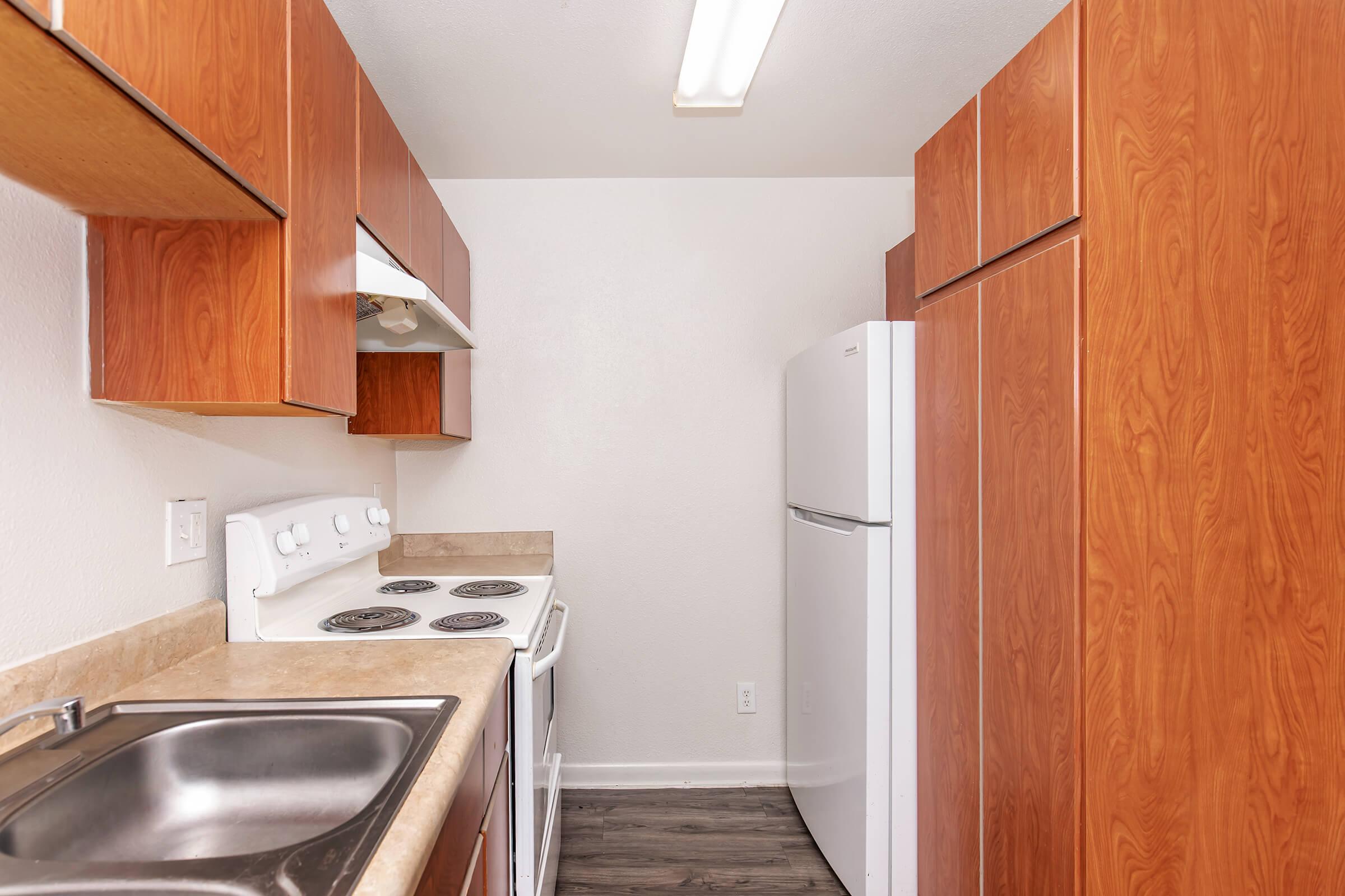 A modern kitchen featuring wooden cabinets, a white stove with four burners, a white refrigerator, and a stainless steel sink. The walls are painted light, and the flooring is dark, creating a warm, inviting atmosphere. The layout is compact and functional, suitable for cooking and meal preparation.