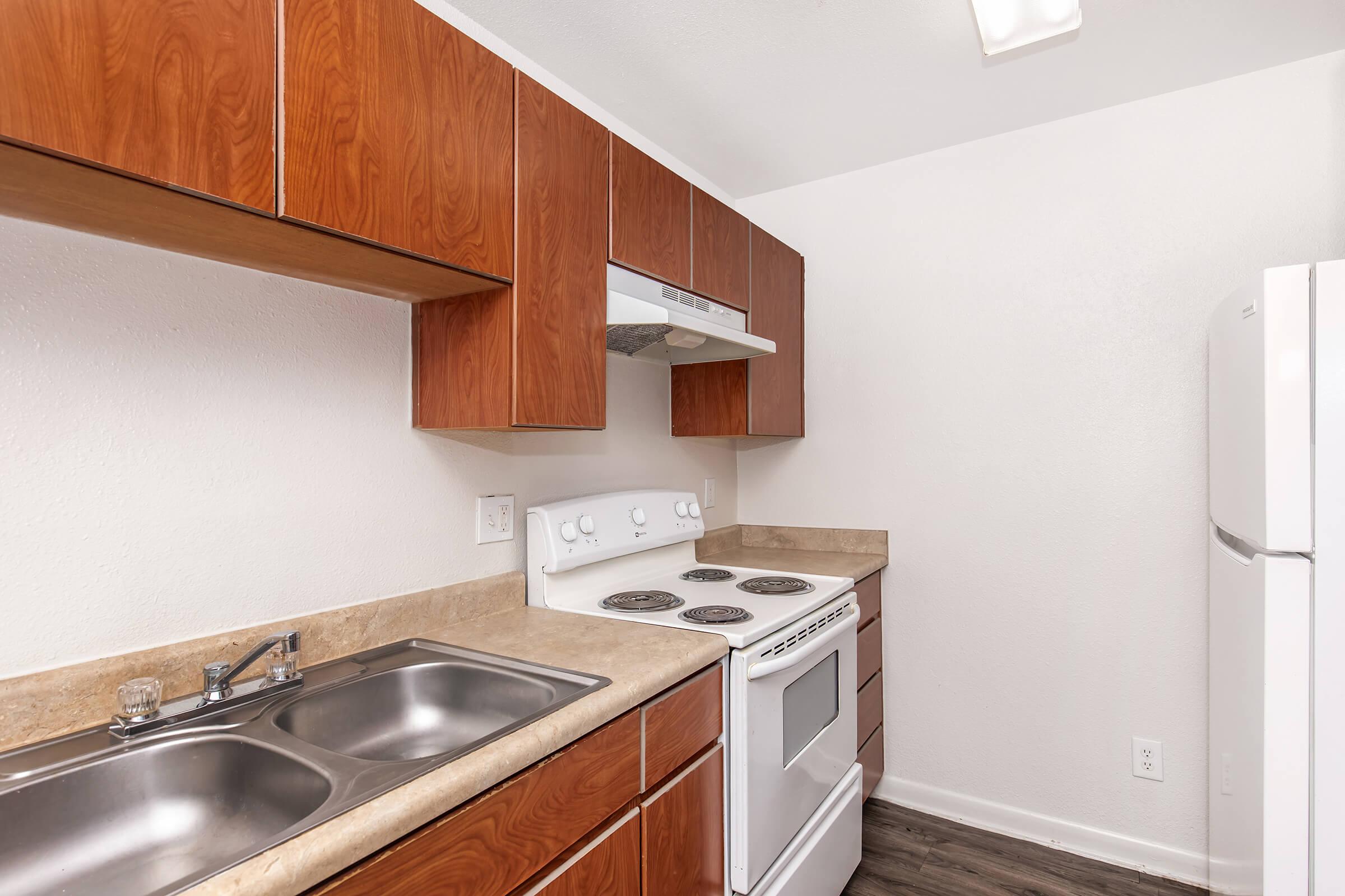 A modern kitchen featuring wooden cabinets, a white stove and oven, a range hood, and a double sink. The countertop is beige, and the flooring is dark. A white refrigerator stands against the wall. The overall color scheme is neutral with a clean, contemporary look.